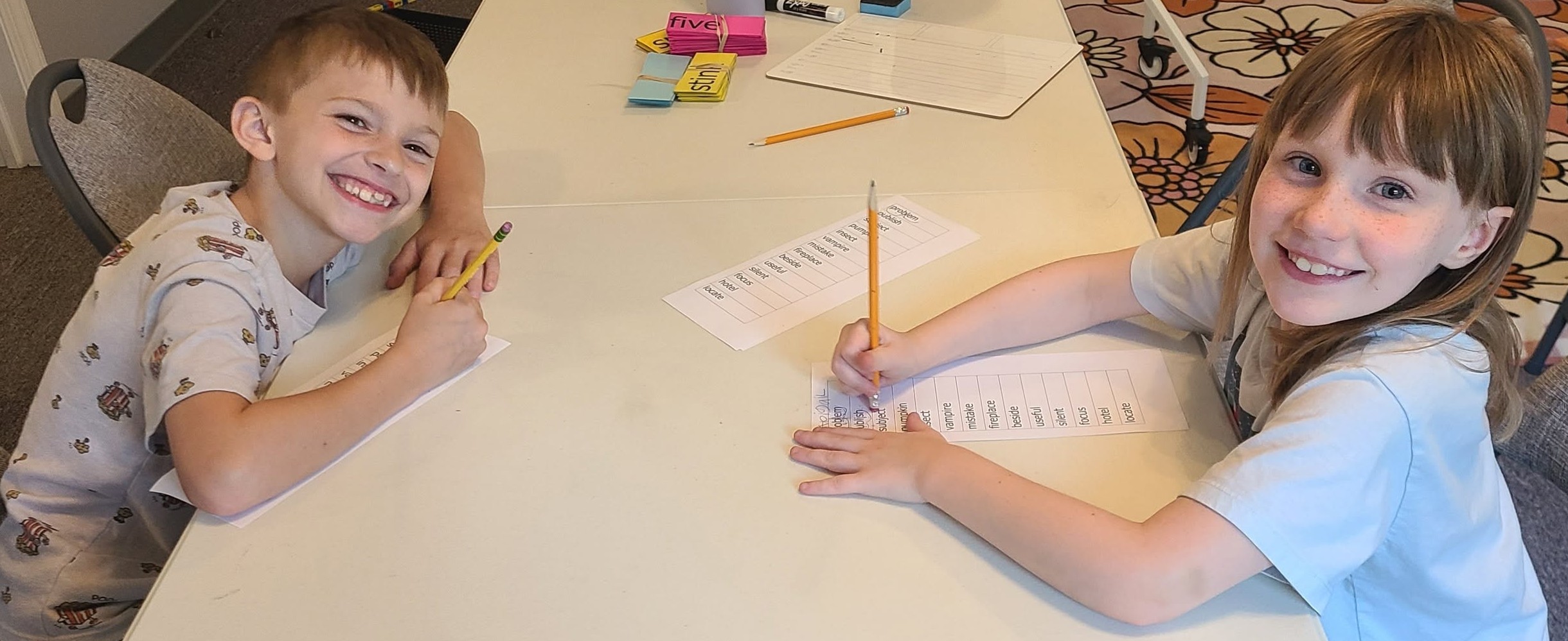 Two children sitting at a table, smiling, with papers and pencils, engaging in an activity.