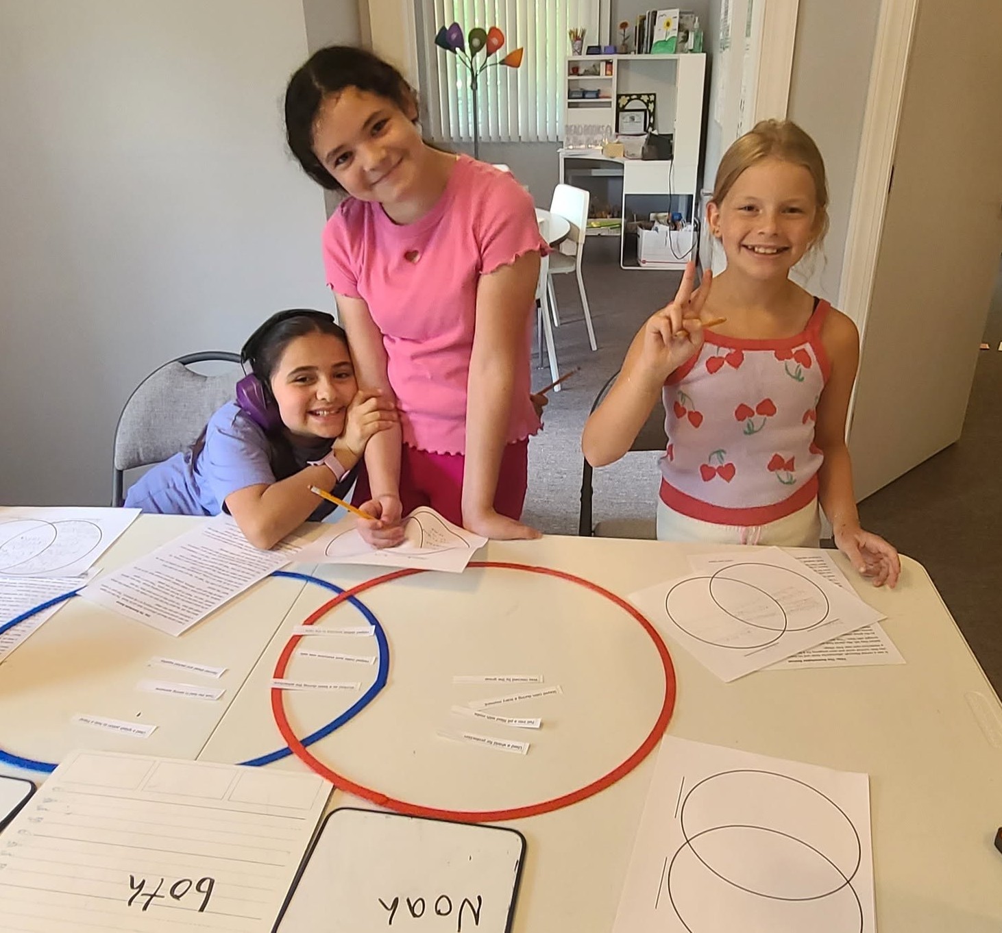 Three smiling children at a table with papers and circles, making peace signs and leaning towards each other.