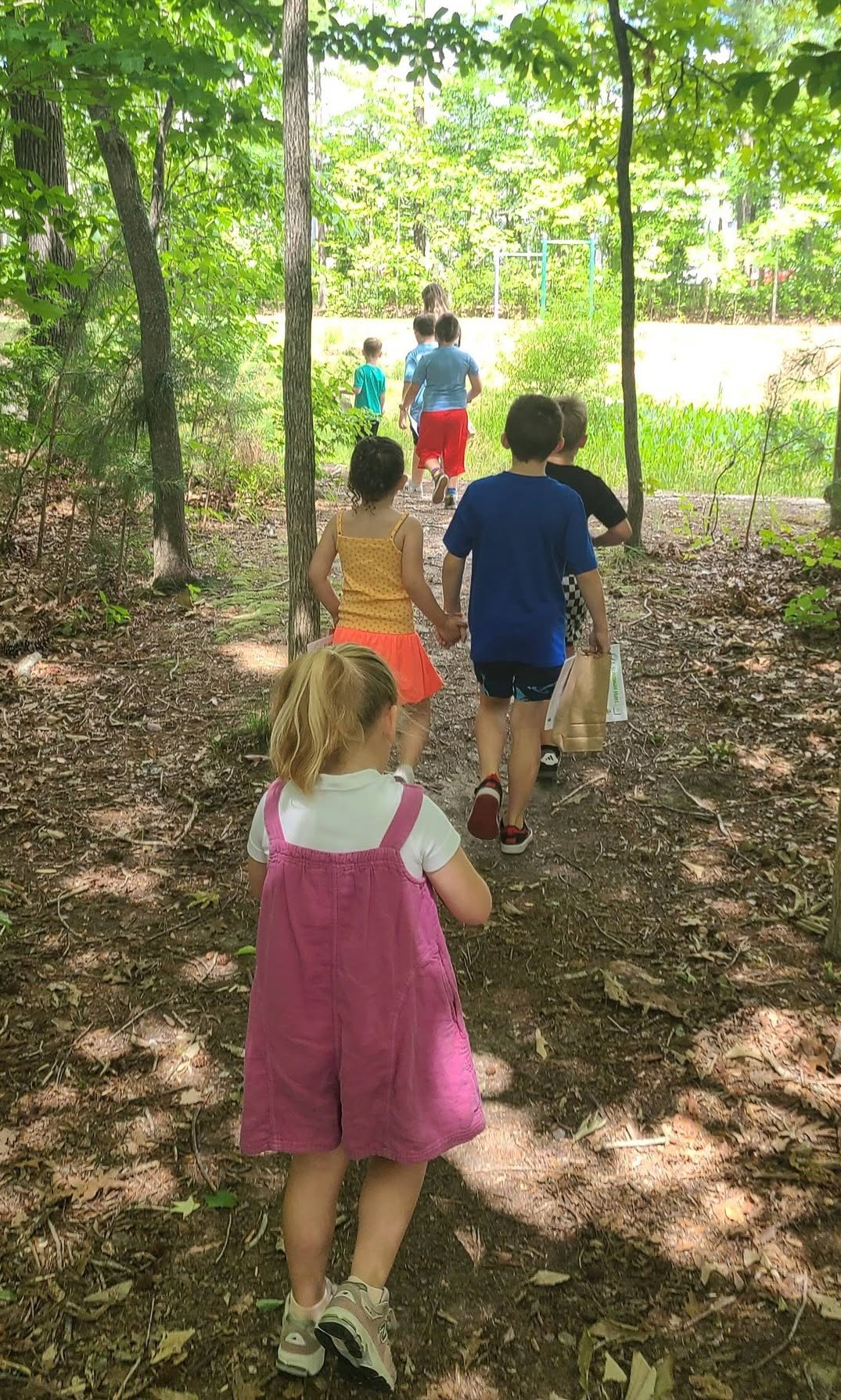 Children walking on a dirt path in a wooded area with sunlight filtering through trees.