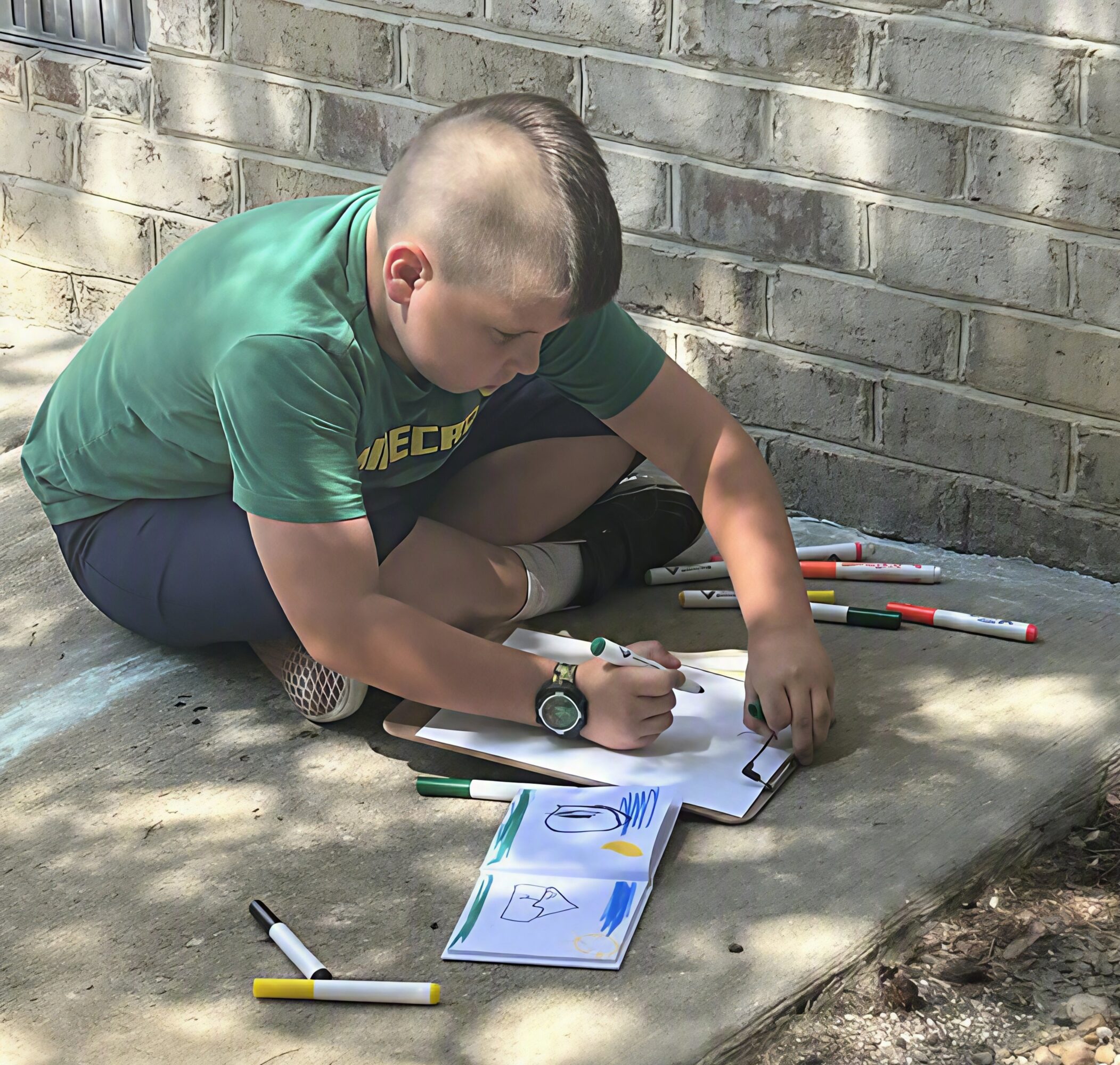 Child sitting on the ground drawing with markers on paper, next to a brick wall, with markers scattered around.