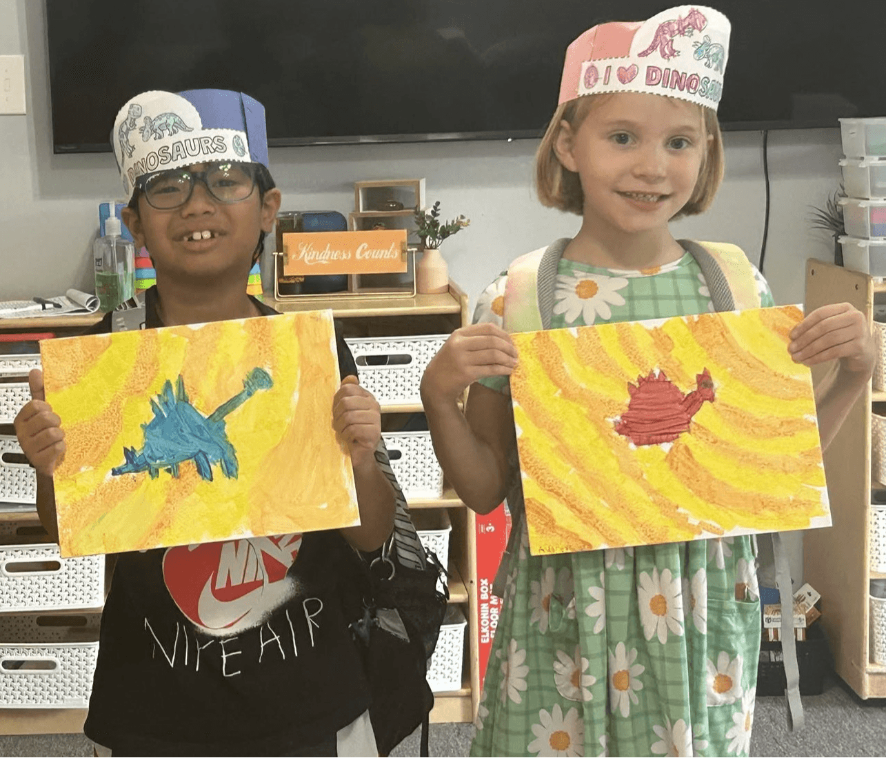 Two children wearing birthday hats hold colorful paintings of animals, smiling in a classroom.