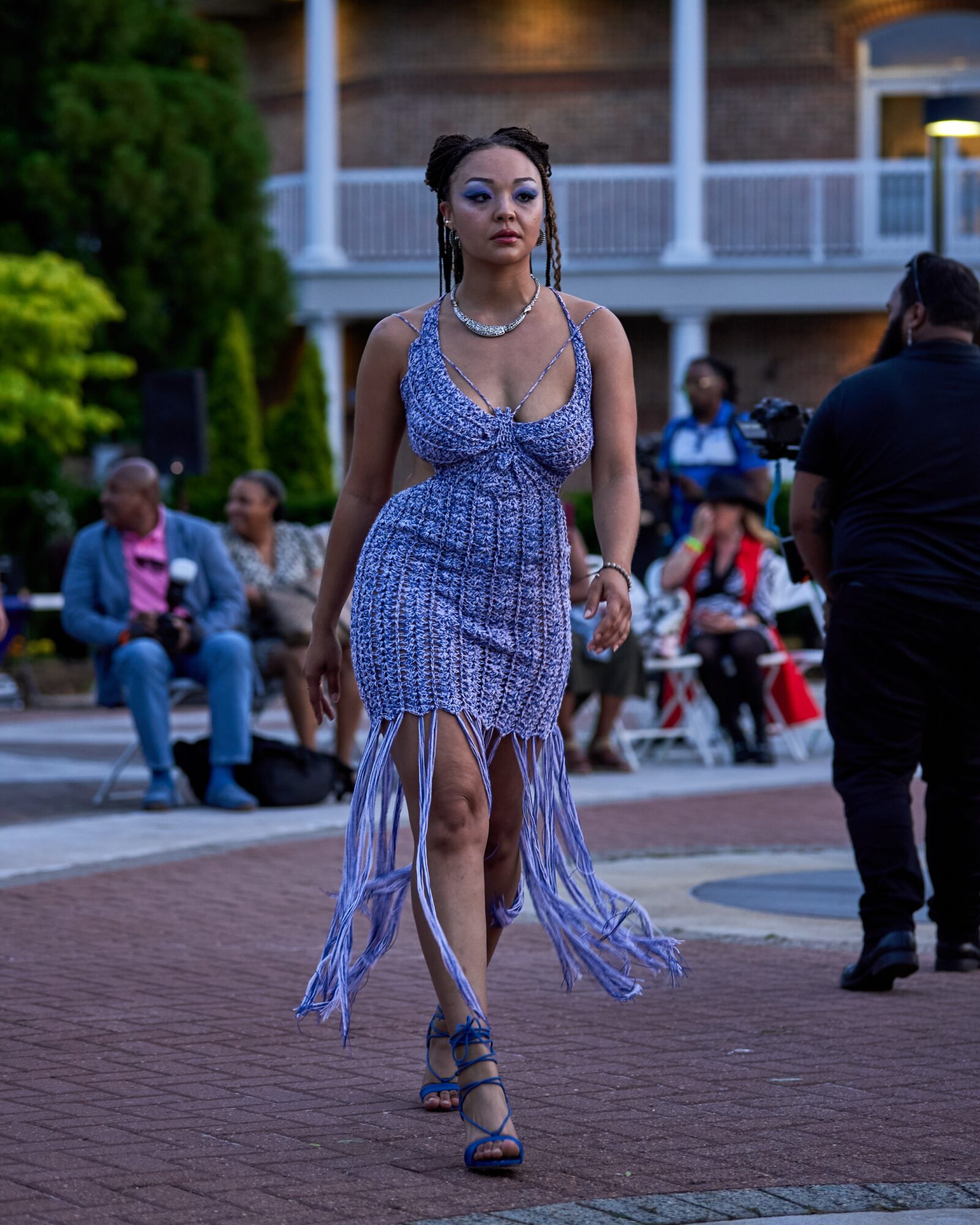 Woman in a blue dress walking outdoors with people seated in the background.