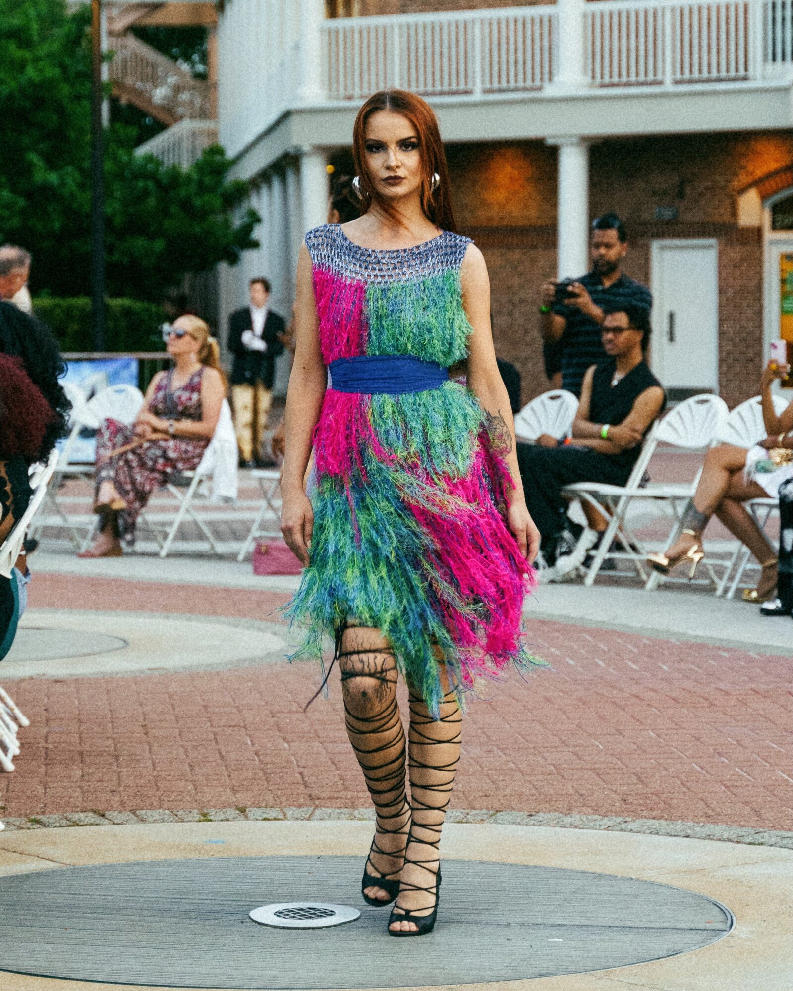 Woman walking on a circular platform in colorful dress and striped tights, with people seated and standing in background.