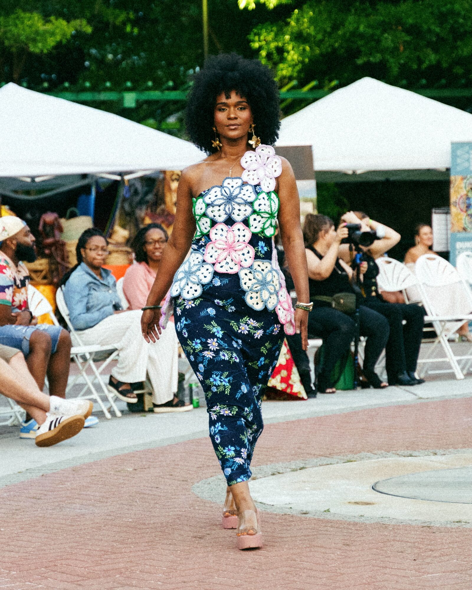 Woman walking outdoors in colorful floral outfit with audience and tents in background.
