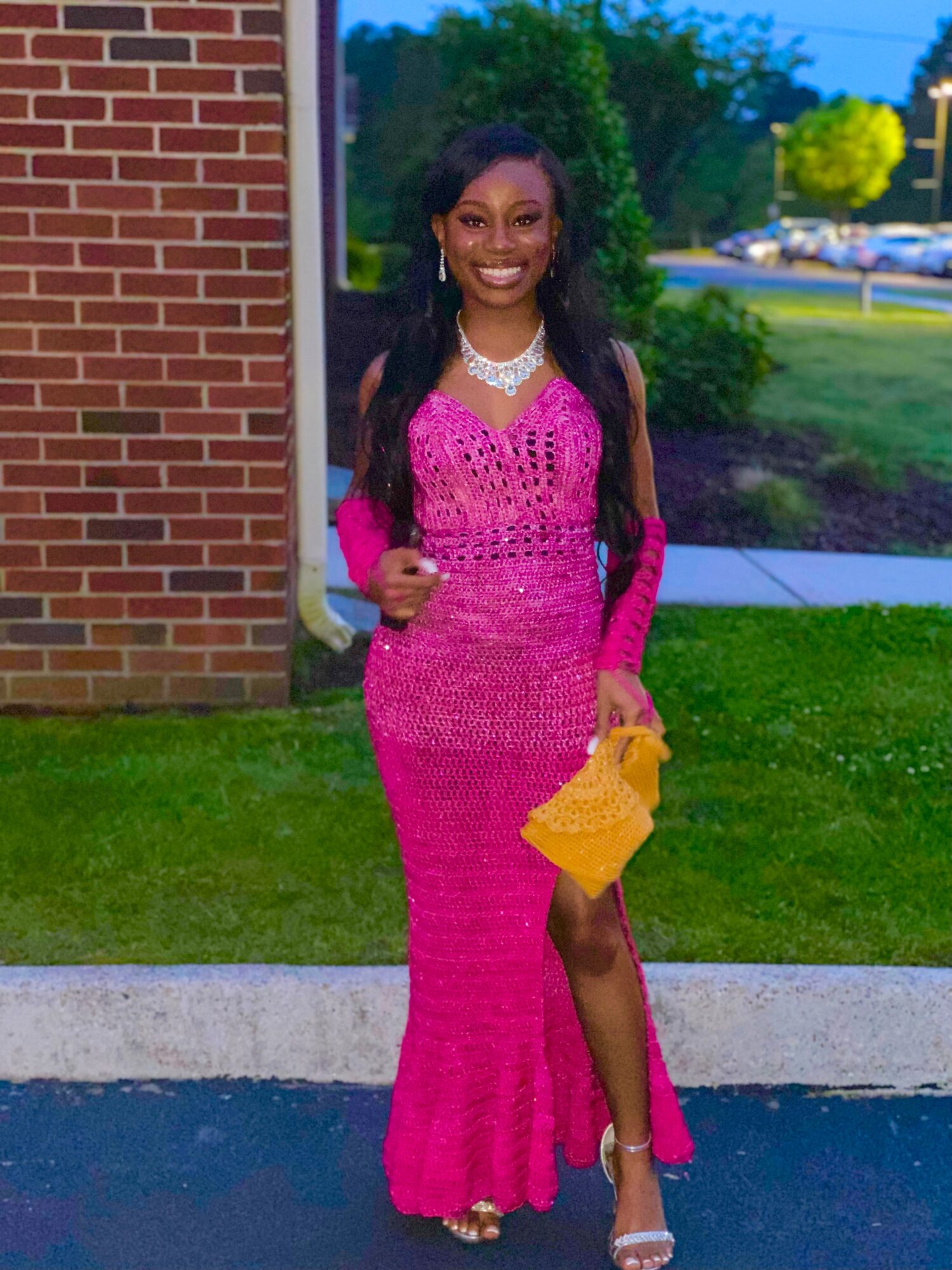 Young woman in a pink gown holding a yellow clutch, standing outdoors near a brick building and greenery.