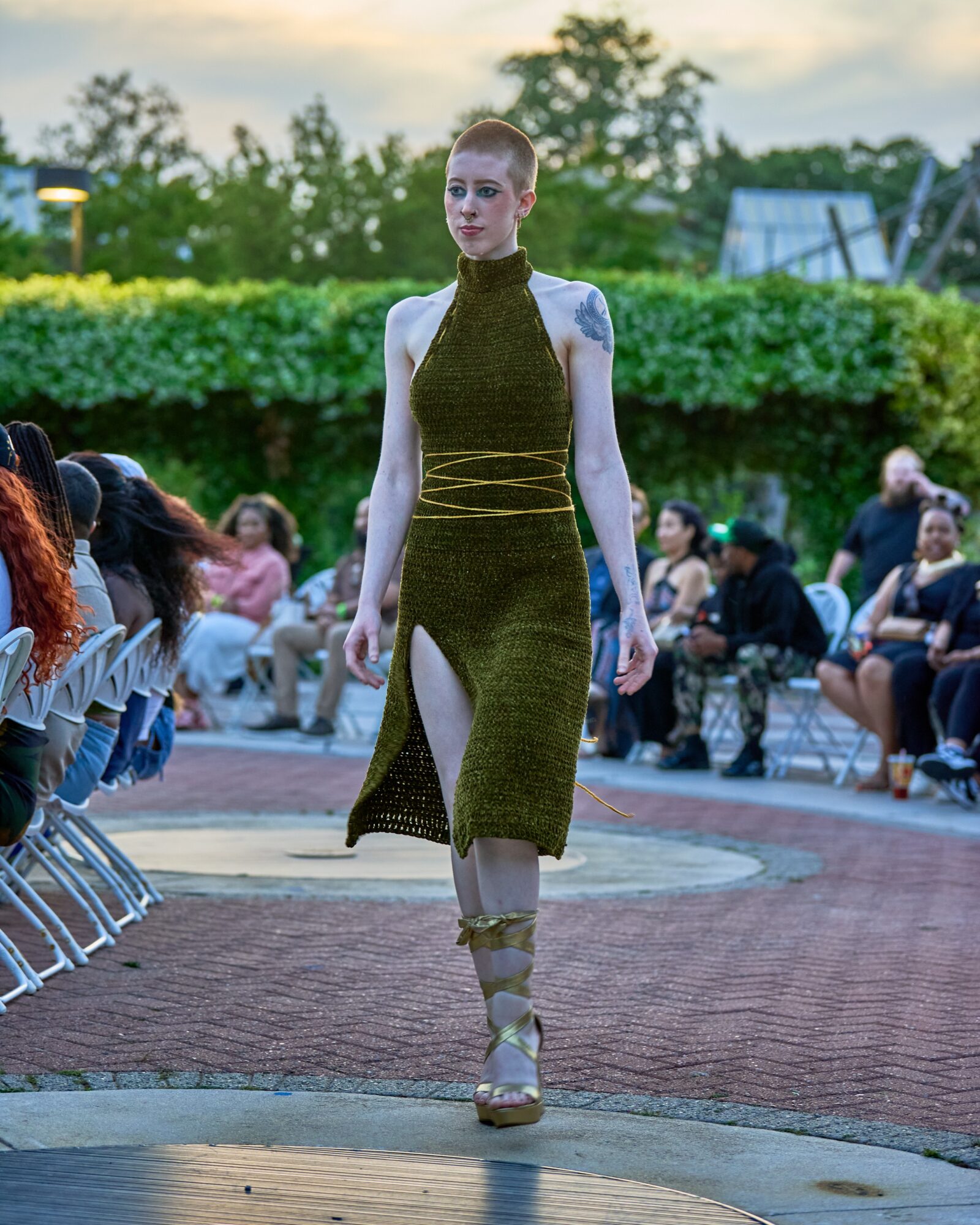 A woman walks on an outdoor runway during a fashion show, with an audience seated on both sides, trees, and a building in the background.