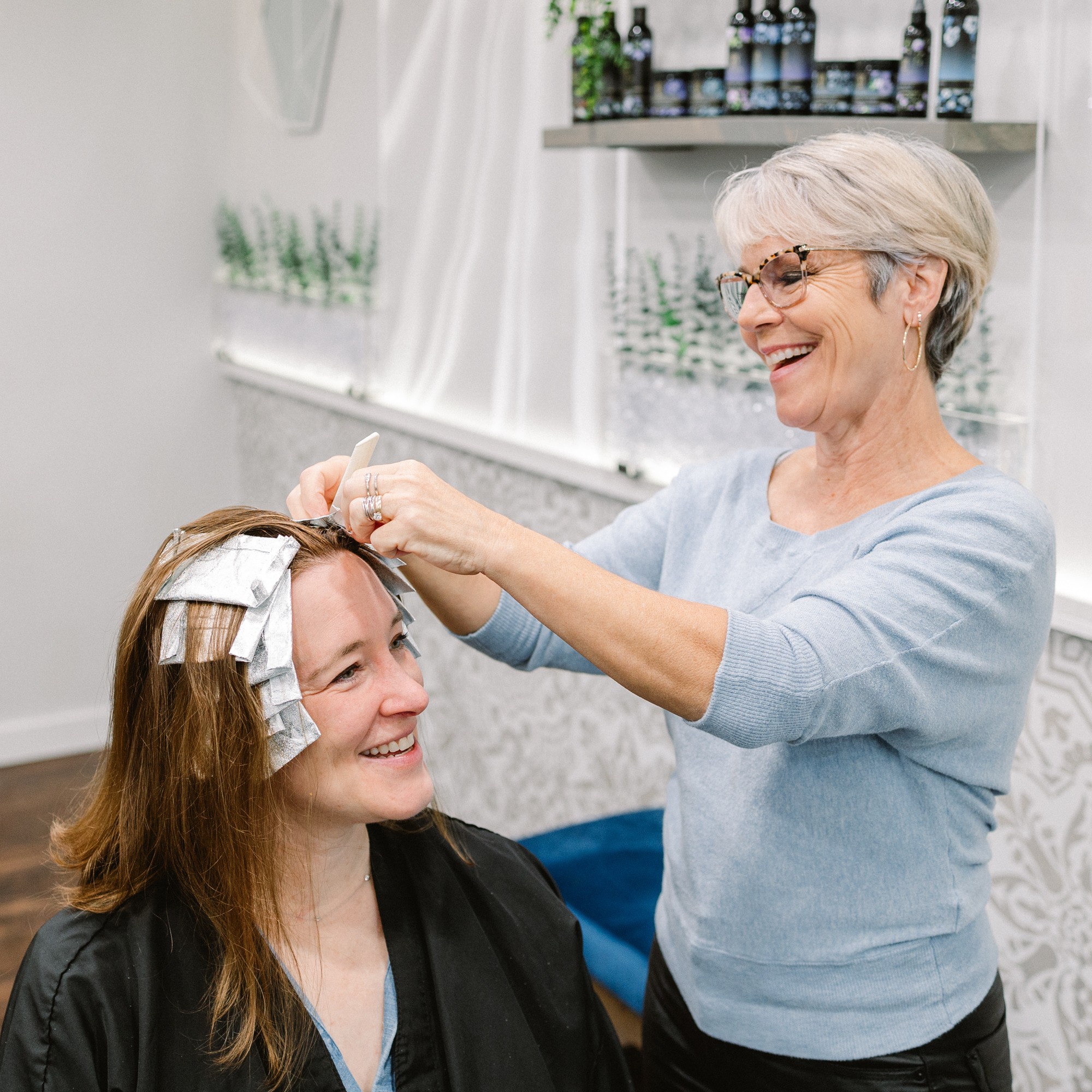Older woman with short gray hair and glasses styles young woman's hair in a salon, both smiling.