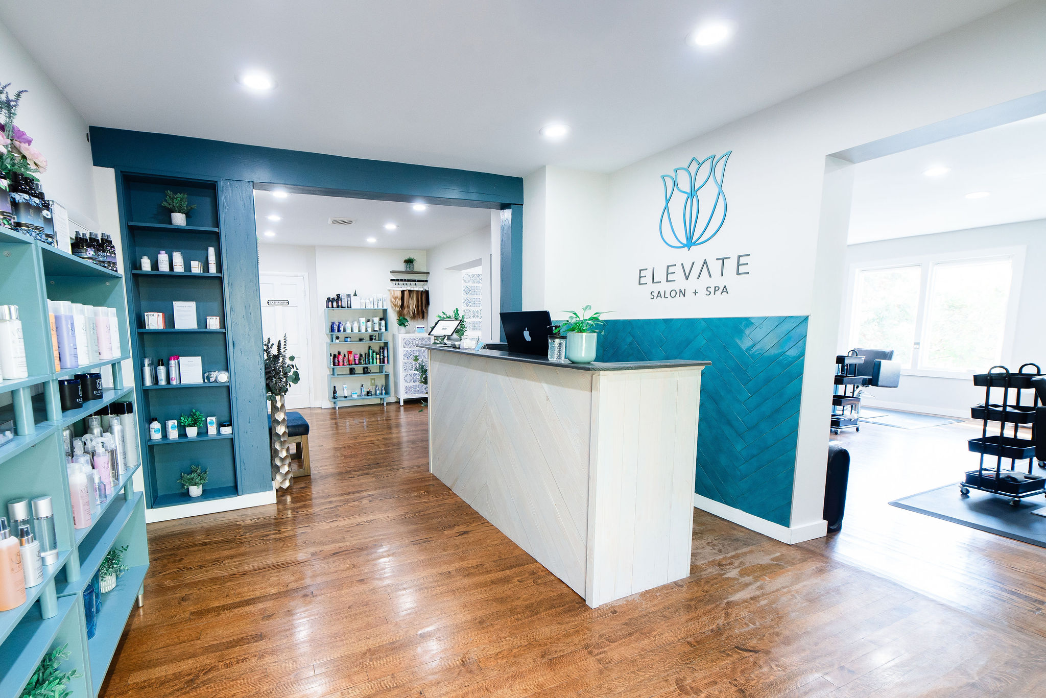 Spa reception area with a white desk, teal accent wall, and shelves with products, wooden flooring, and natural light.