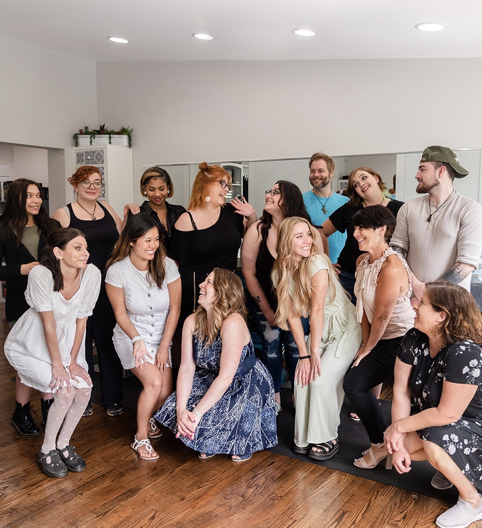 Group of people gathered indoors, some standing and some sitting, in a room with white walls and wooden floor.