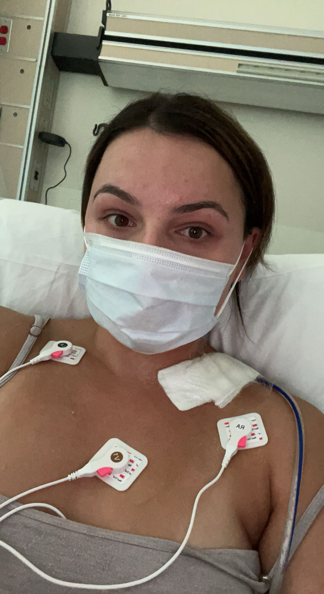 Woman lying in hospital bed wearing a face mask and medical wires attached to her chest.