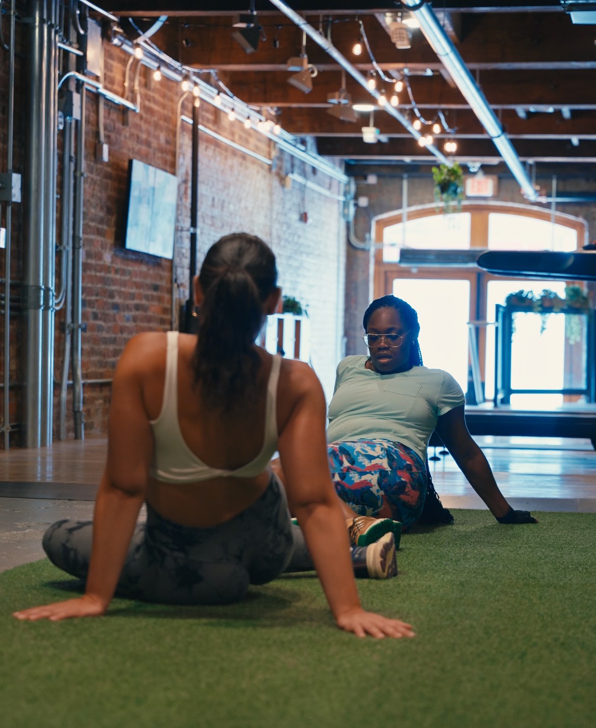 Two women sitting on the floor in a gym with brick walls and large windows, engaging in conversation.