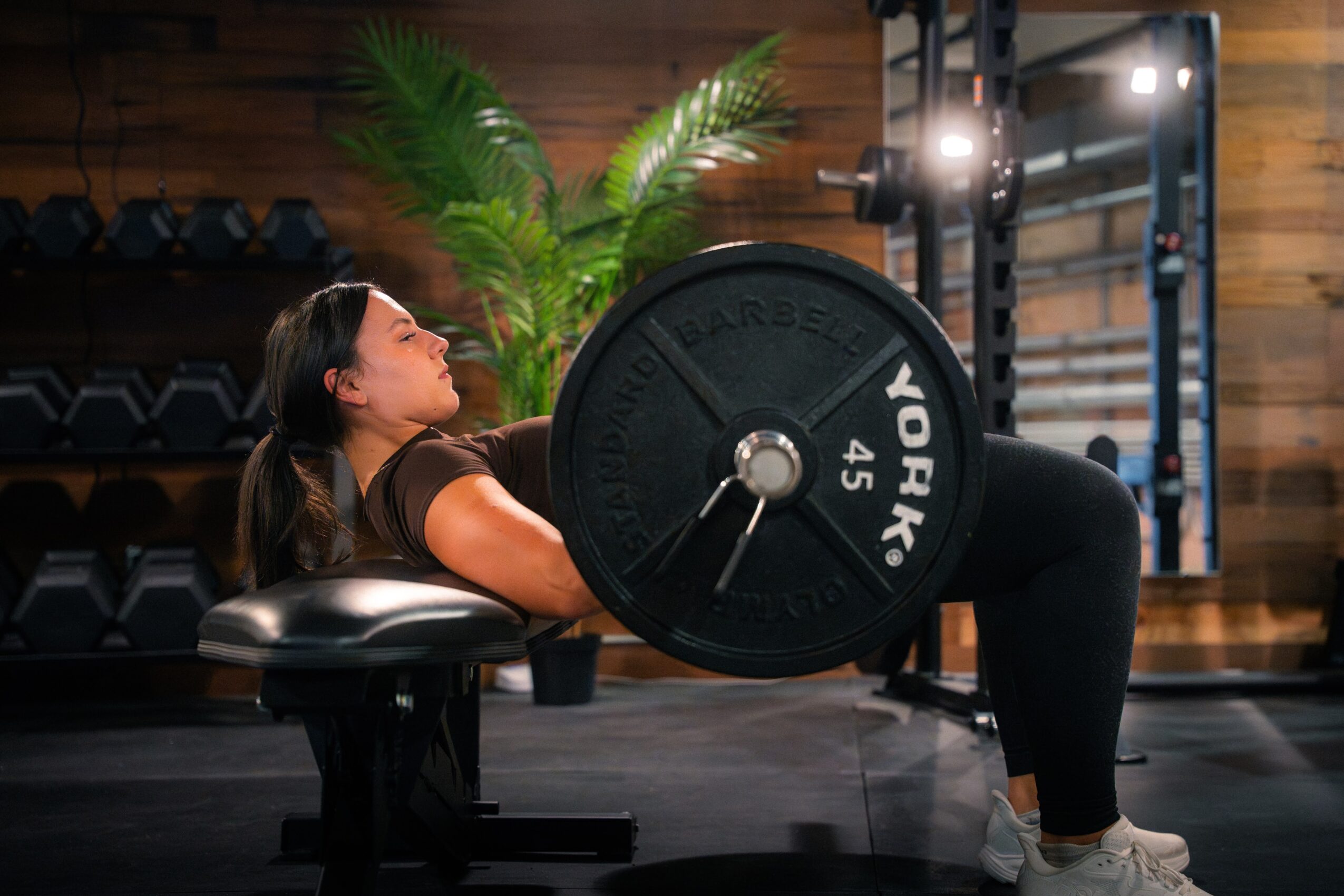Woman lying on a bench lifting a barbell with weights in a gym, with wooden walls and plants in the background.