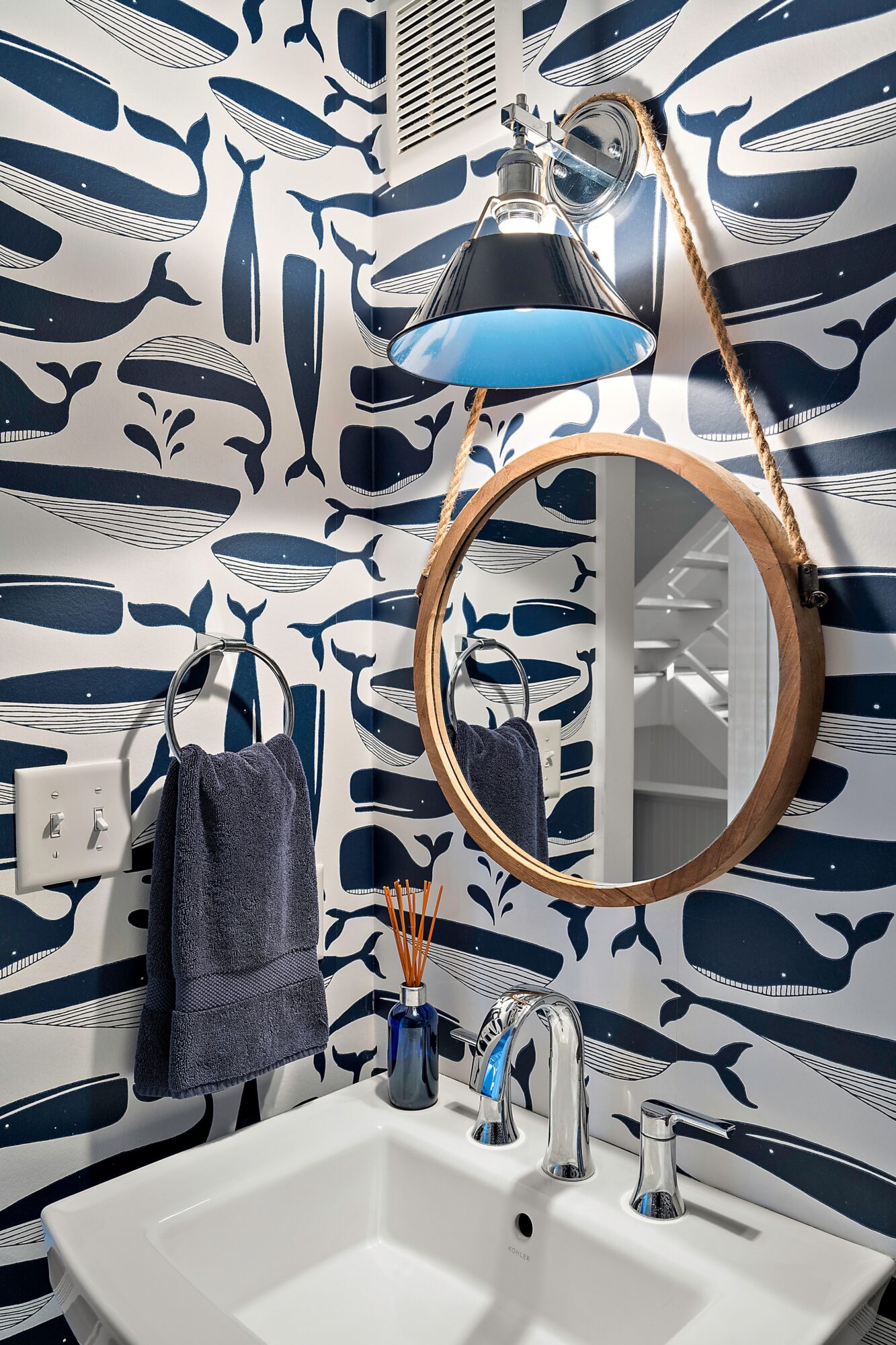 Bathroom mirror and sink with patterned wallpaper, towel, and toiletries, illuminated by a wall-mounted light fixture.