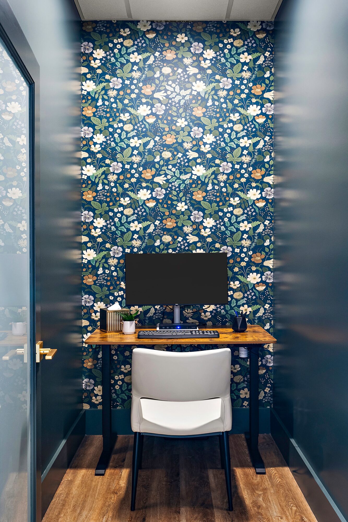 Small workspace with floral wallpaper, desk, and white chair, enclosed by glass walls.