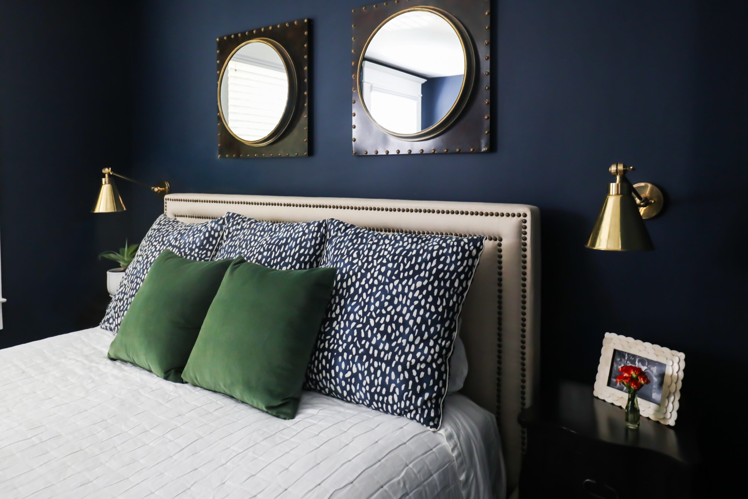 Bedroom with dark blue walls, bed with patterned and green pillows, two wall mirrors, and a nightstand with a framed photo.