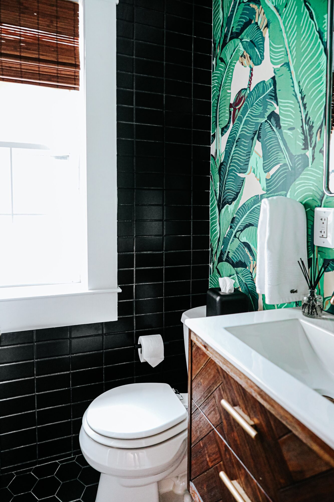 Bathroom with black tiled wall, tropical leaf wallpaper, white sink, and toilet, with a window and wooden blinds.