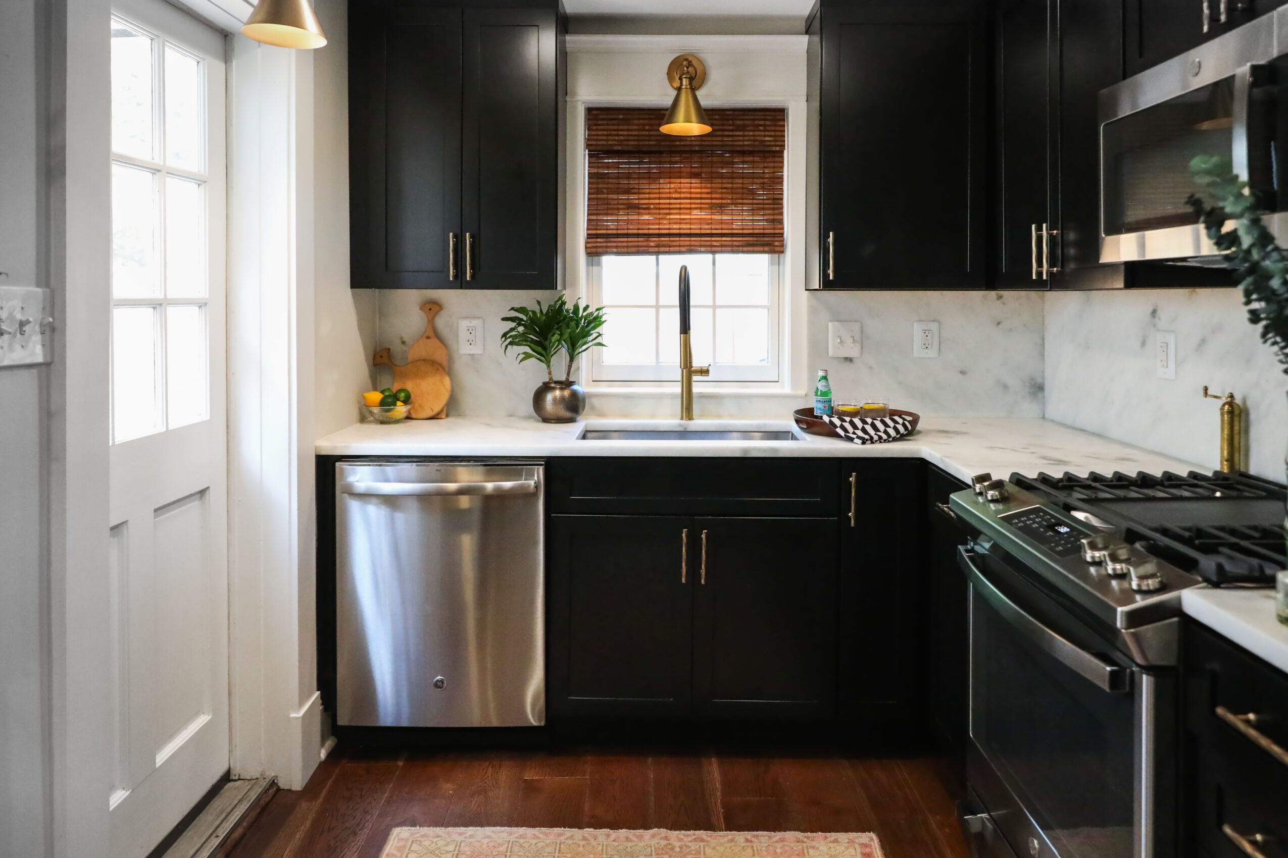 Kitchen with black cabinets, white marble backsplash, stainless steel dishwasher, and a window above the sink, with a wooden floor.