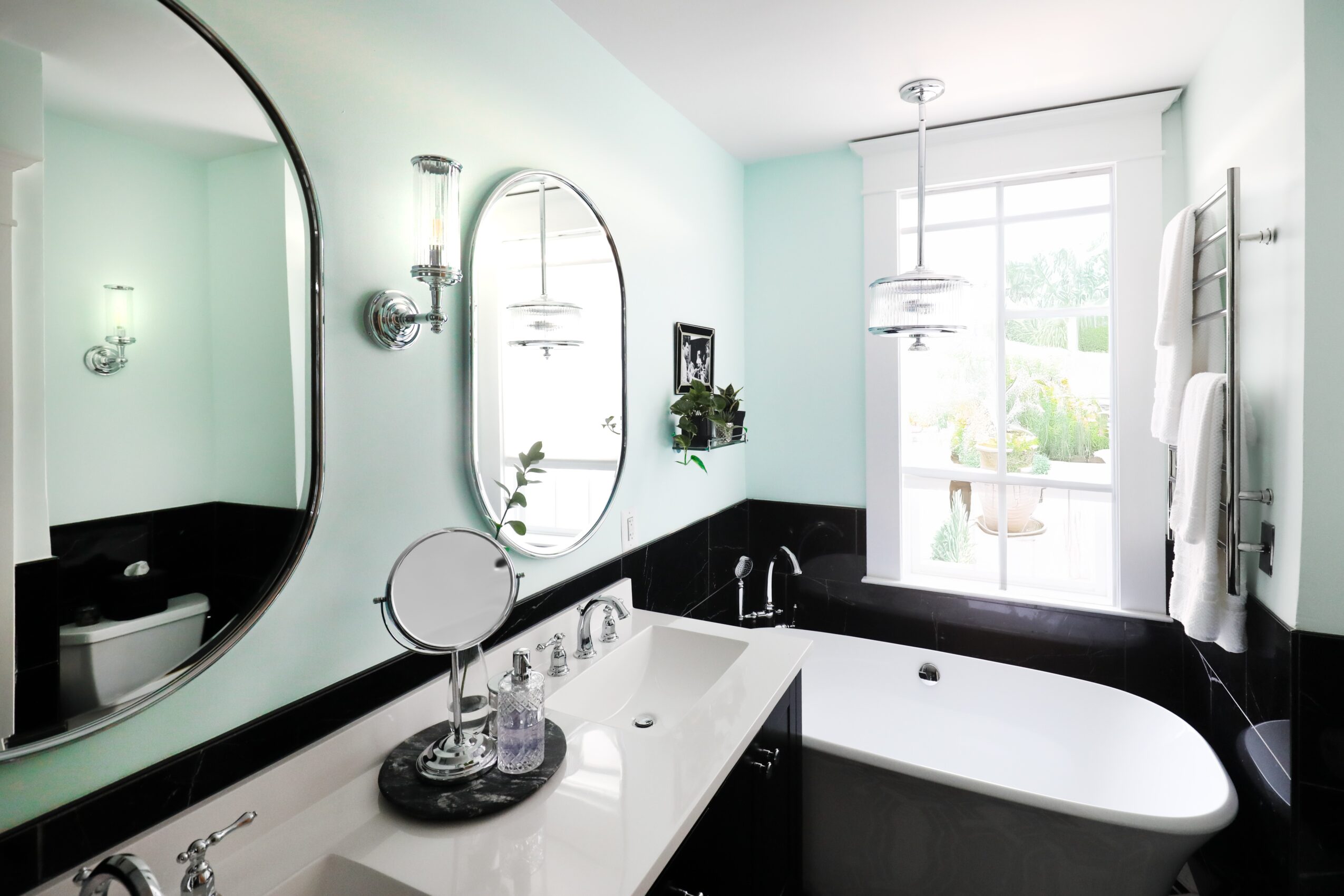 Bathroom with oval mirror, bathtub, and window, decorated with plants and toiletries, in a bright space with light-colored walls.