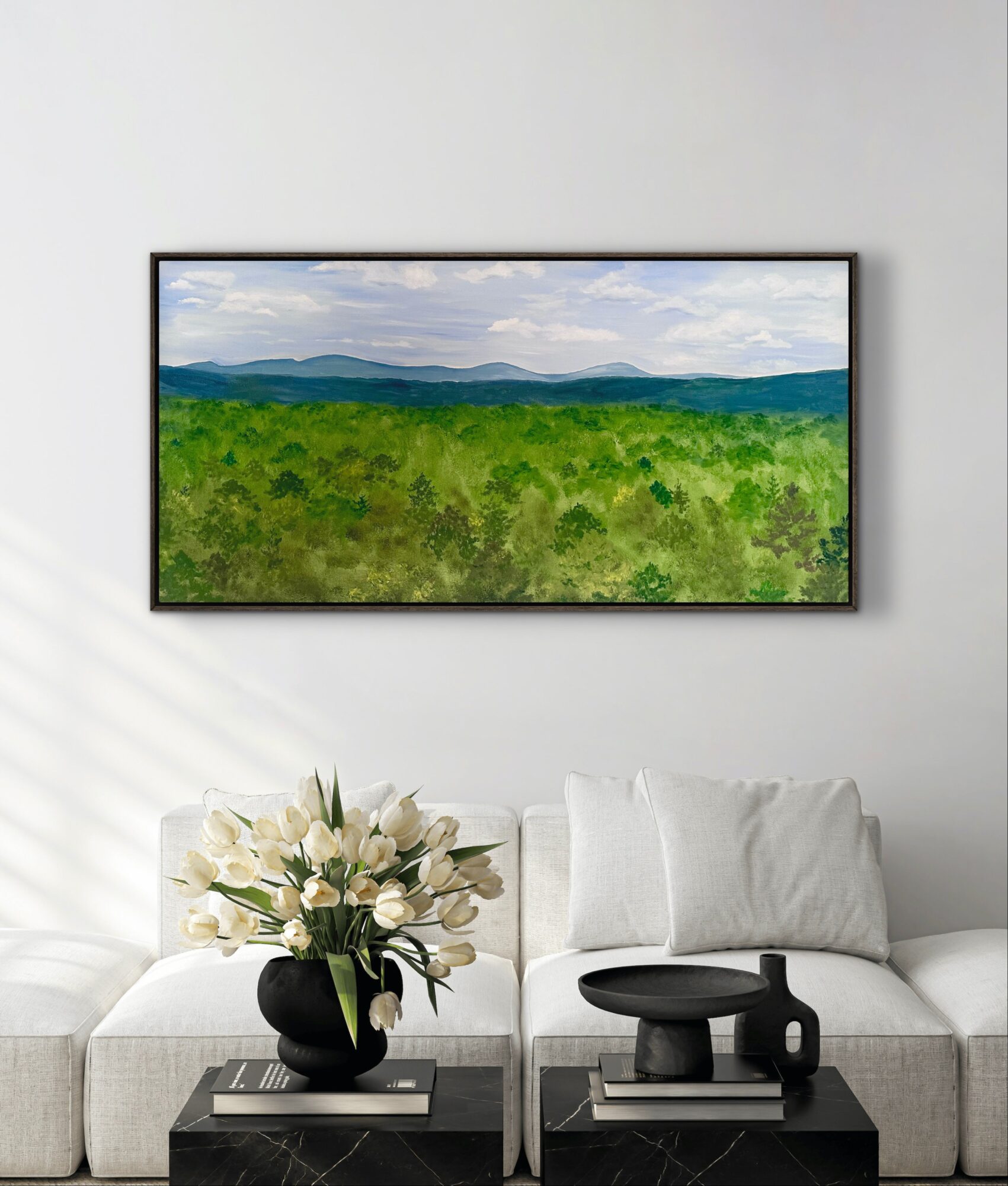 Landscape painting of mountains and green fields hanging above a white sofa with a flower arrangement and decorative objects.