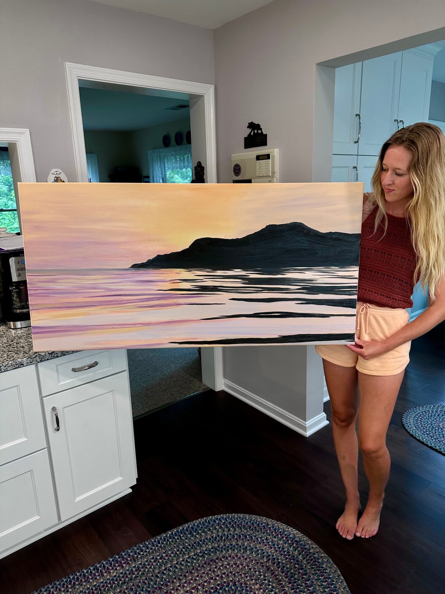 Young woman with long blonde hair holding a landscape painting of a mountain and water, standing in a room with white cabinets.