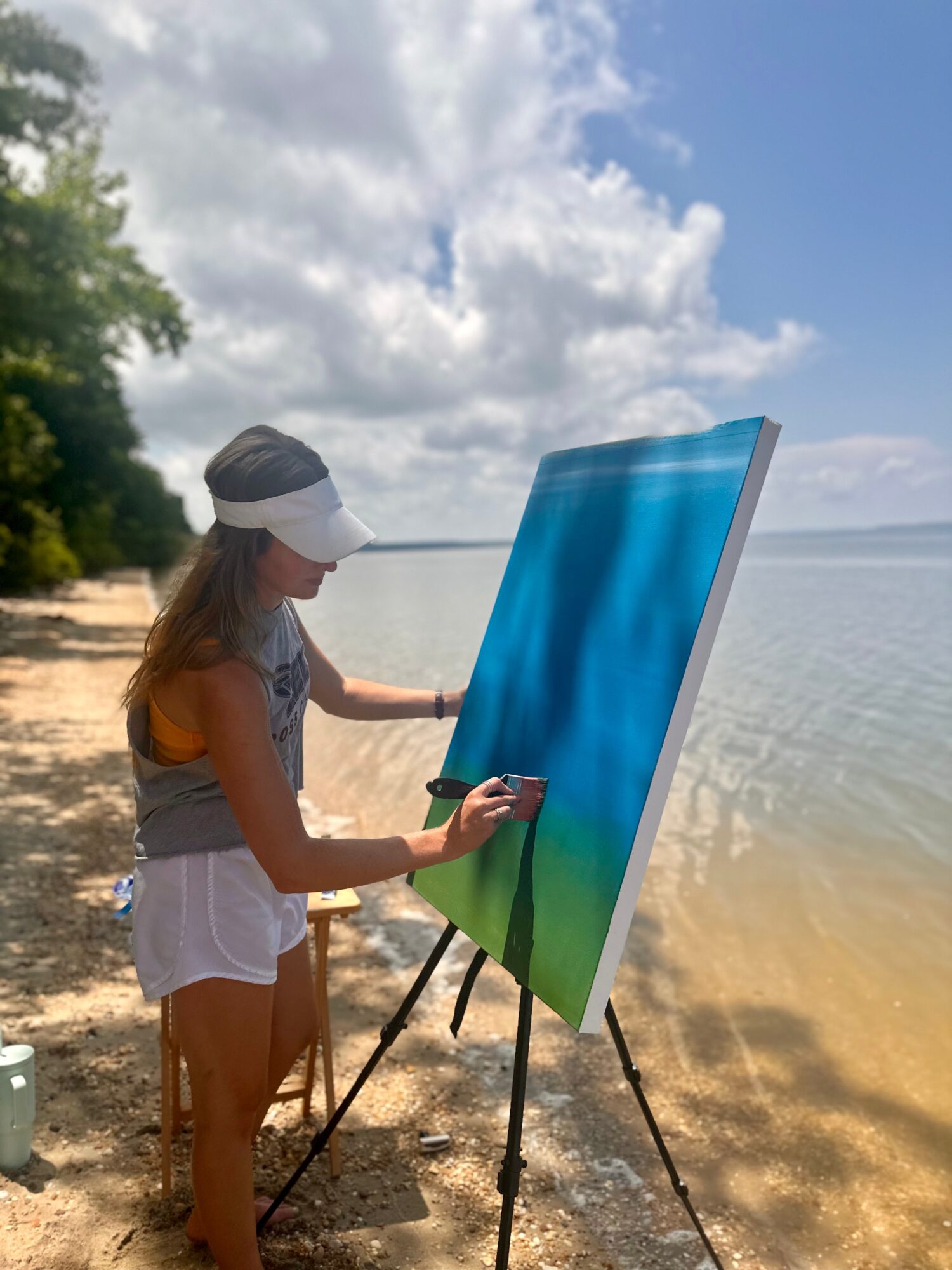 Child painting on canvas outdoors by water, wearing a visor and casual clothing, with trees and cloudy sky in background.