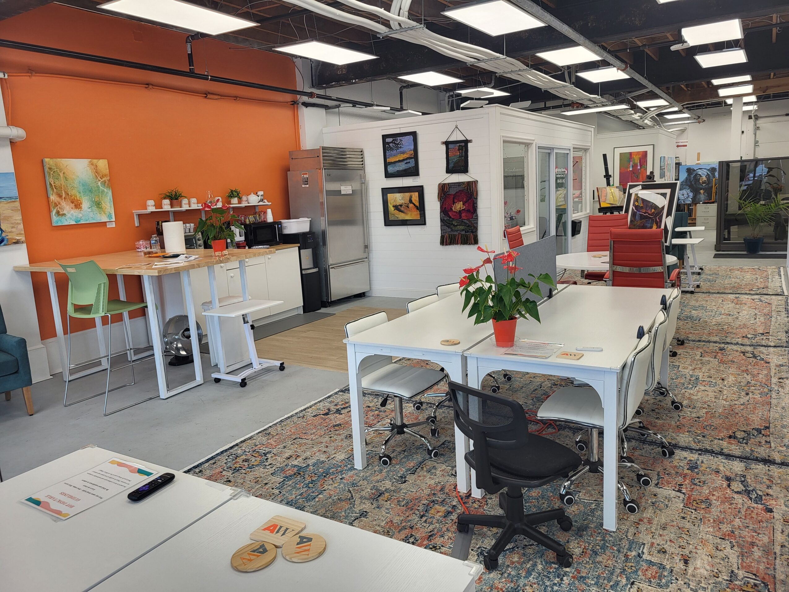 Open office space with tables, chairs, a potted plant, and artwork on the walls, with a colorful rug on the floor.