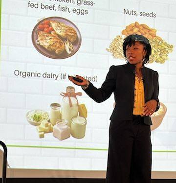 Woman in black suit presenting in front of a food chart with images of chicken, nuts, and dairy products.