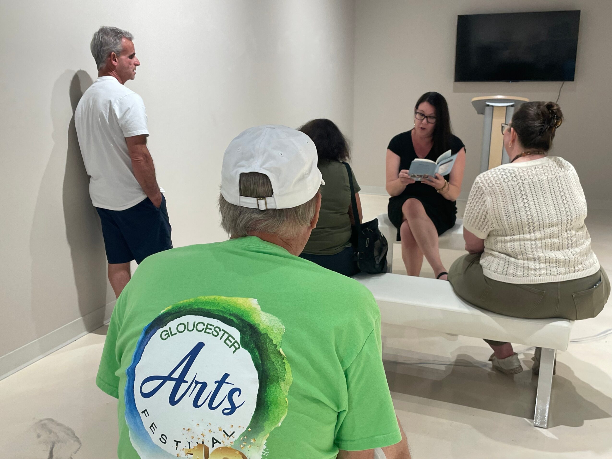 Group of five people in a room, one person reading aloud, others seated and listening, TV on wall, plain white walls.