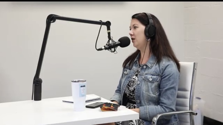 Woman wearing headphones speaking into a microphone in a recording studio, sitting at a white table with a notebook and a drink.