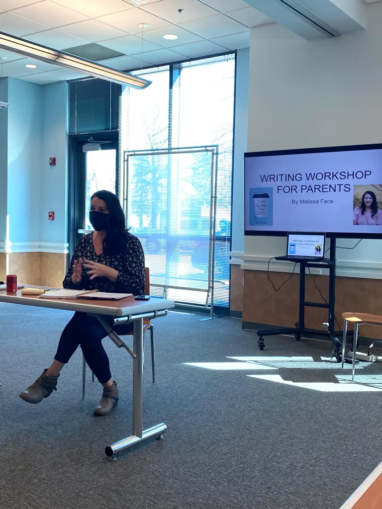 Woman wearing a mask sitting at a table in a bright room with large windows, a screen displaying a writing workshop for parents, and a small table with a laptop.