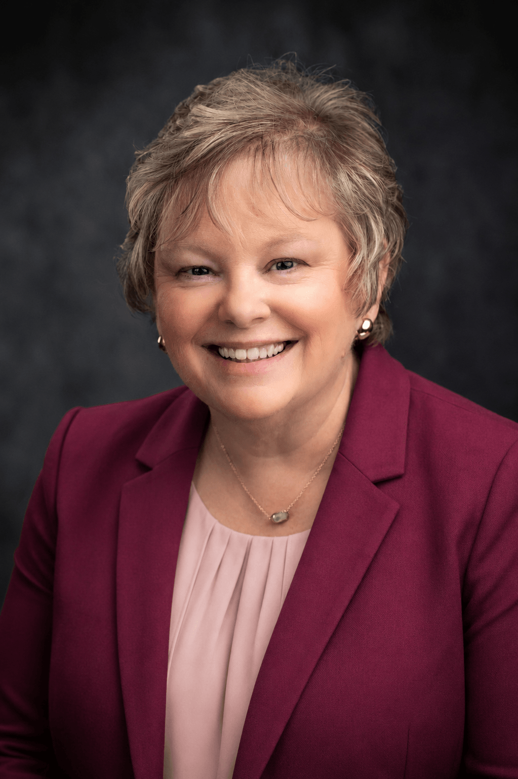 Smiling woman with short hair wearing a maroon blazer and light pink blouse, against a dark background.