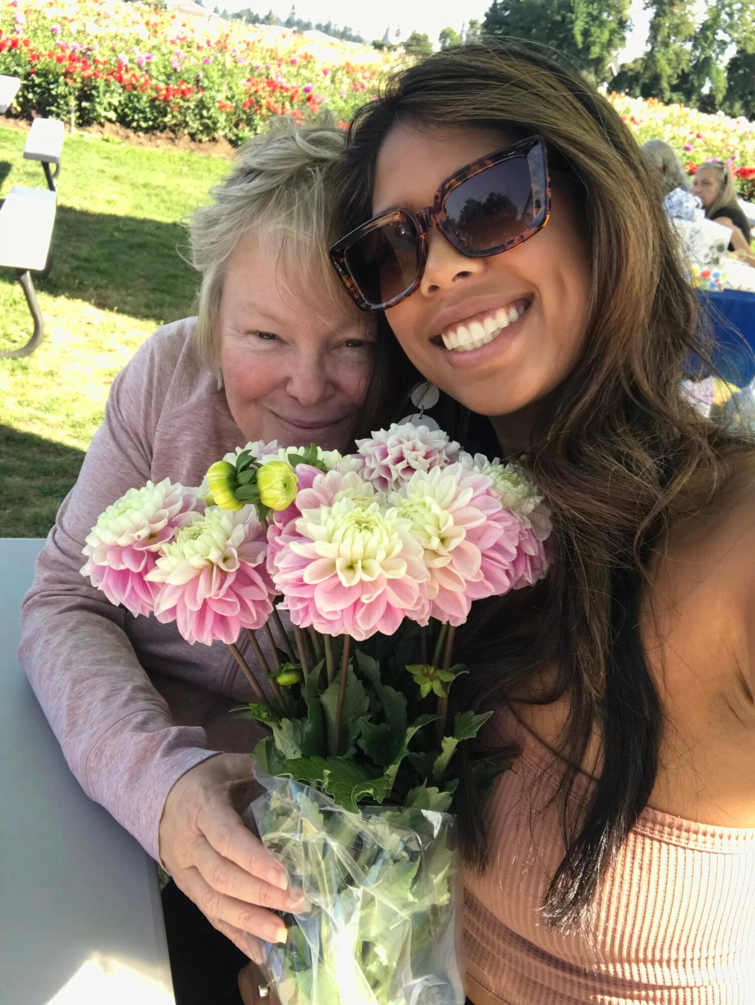 Two women smiling, holding a bouquet of pink and white flowers outdoors on a sunny day.
