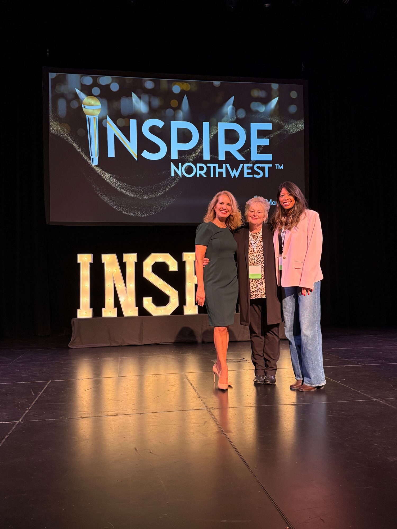 Three people standing on stage in front of a large screen with the word INSPIRE, with a table in front of them.