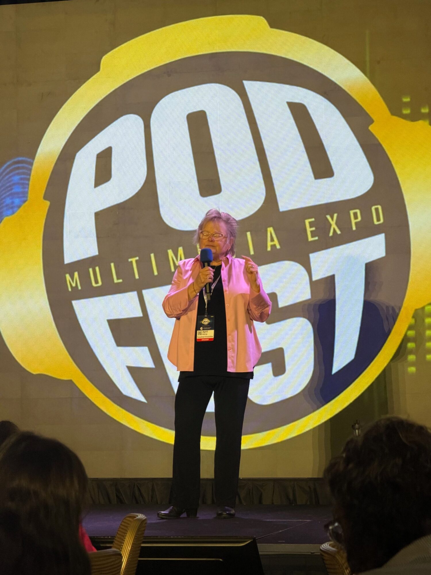 Two women stand on stage in front of a large circular logo with the words 'Quad Expo' and 'Multimedia' visible. One woman holds a microphone.