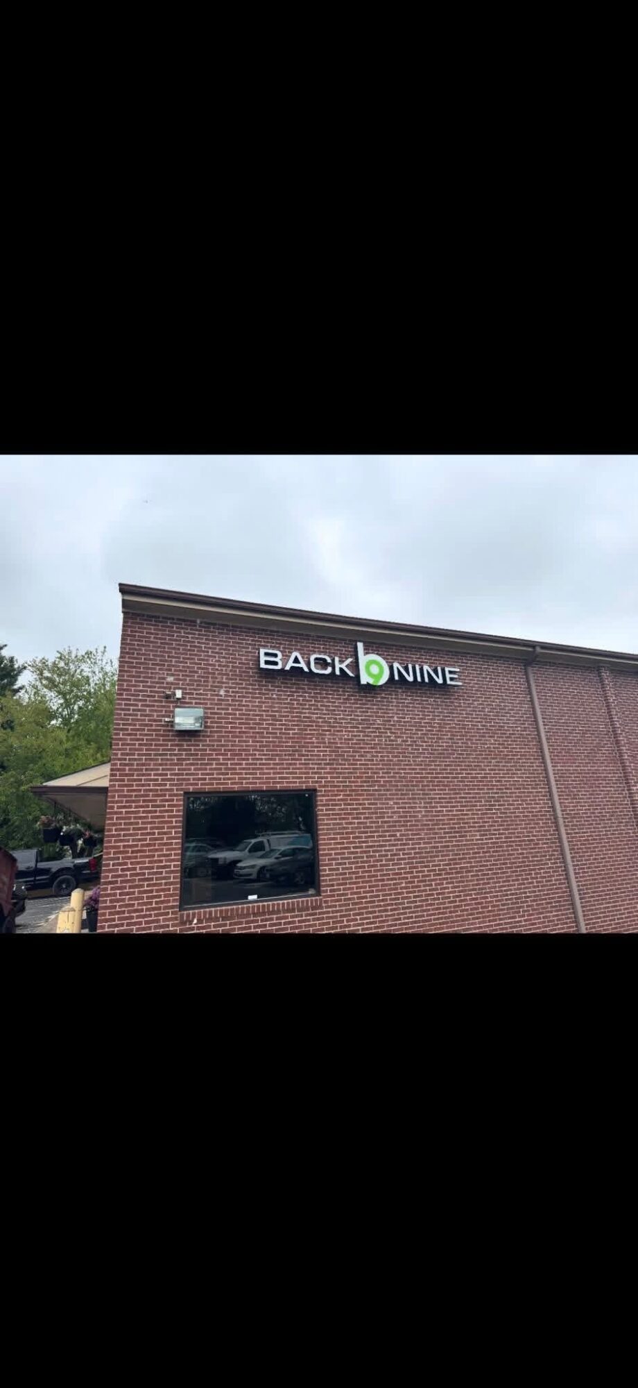 Brick building with a sign reading 'BACK NINE' and a window, outdoors with trees in the background.