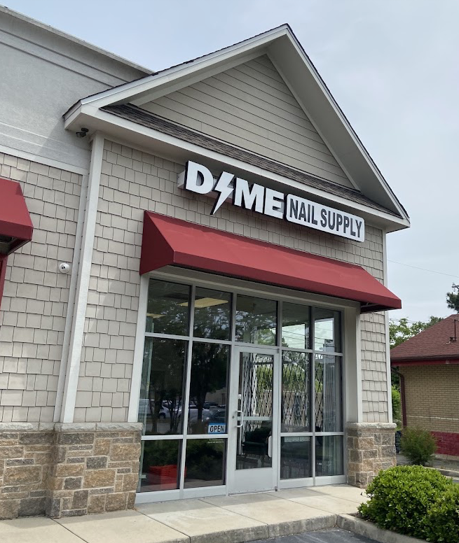 Storefront with a sign reading 'DAME NAIL SUPPLY' above glass doors, red awning, and brick and siding exterior.