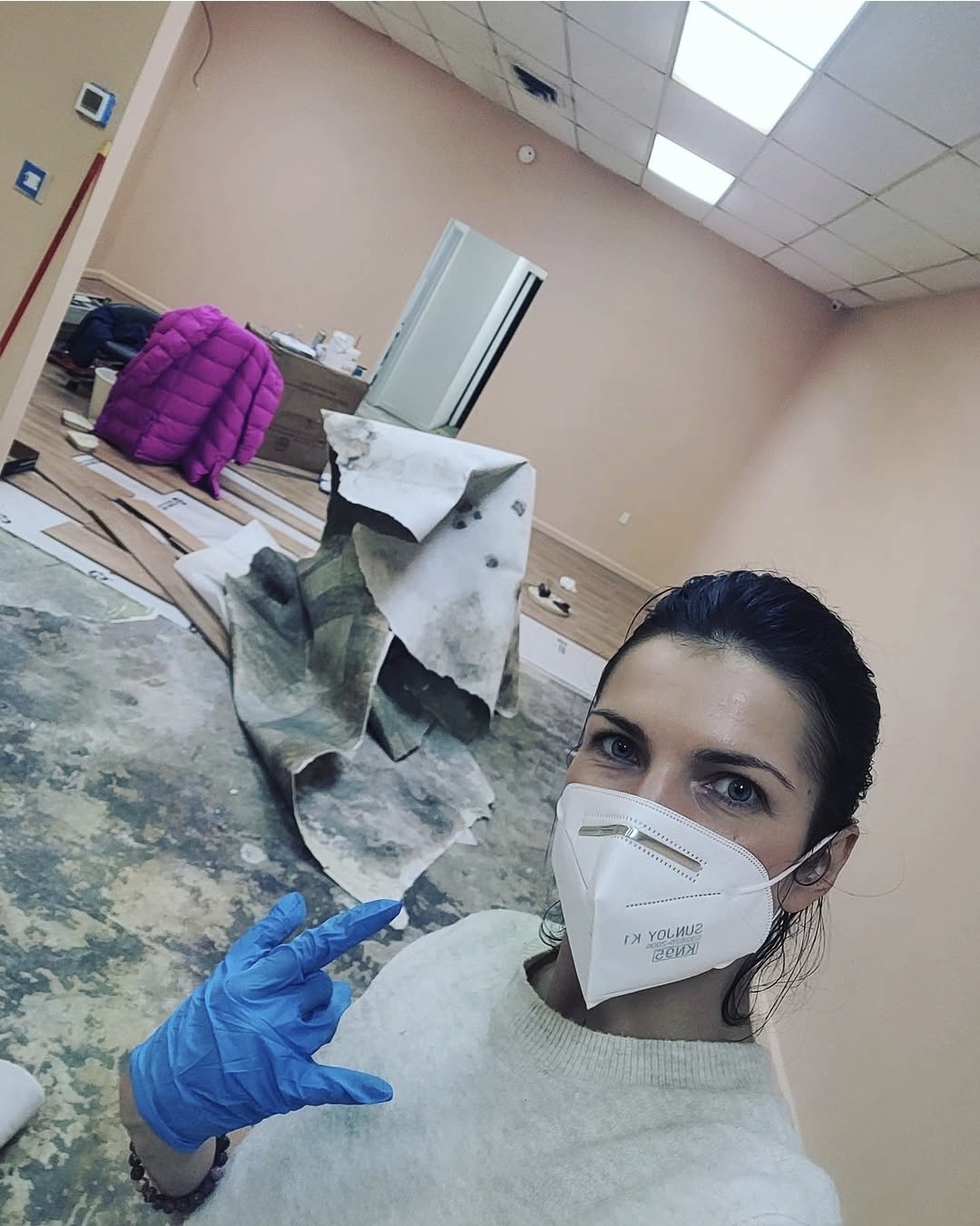 Woman wearing a mask and blue gloves taking a selfie in a room with cleaning supplies and a pink jacket.