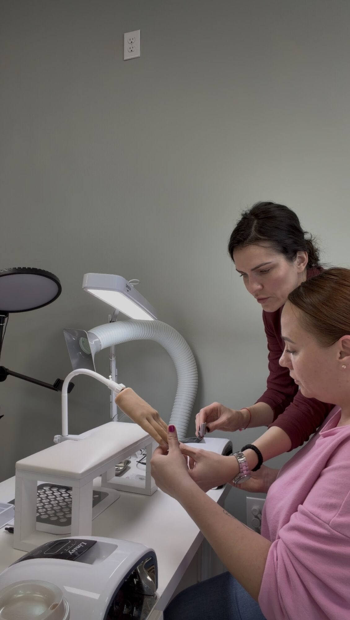 Two women in a clinical setting examine a hand using medical equipment, with a gray wall in the background.