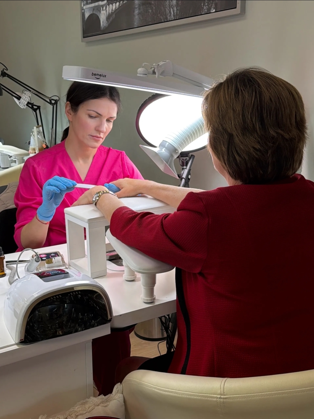 A woman in a pink uniform and blue gloves performs a manicure for a person with short brown hair in a salon. The scene includes manicure tools and a lamp.