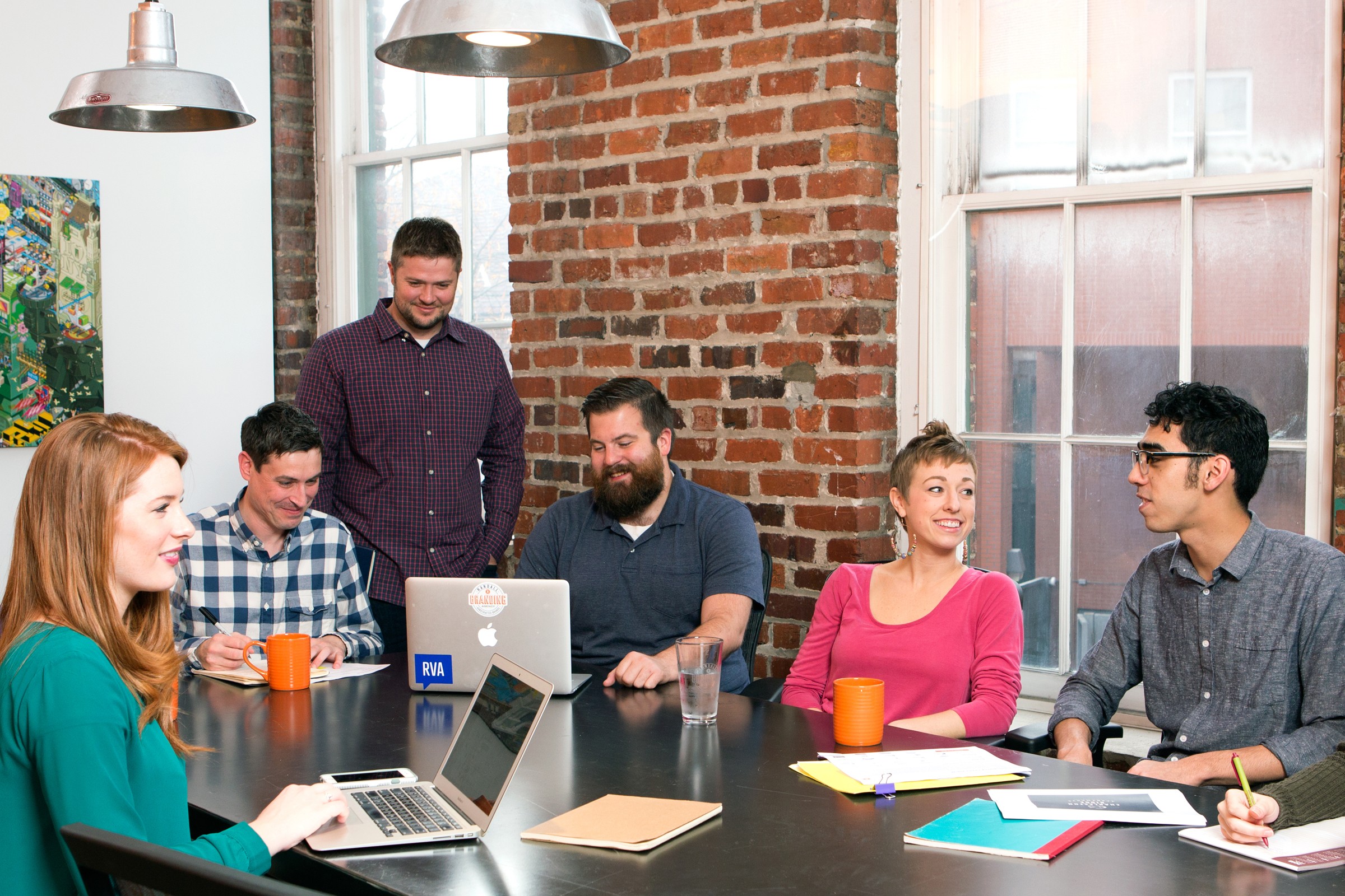 Group of six people sitting around a table in a room with brick wall and large windows, working on laptops and papers.