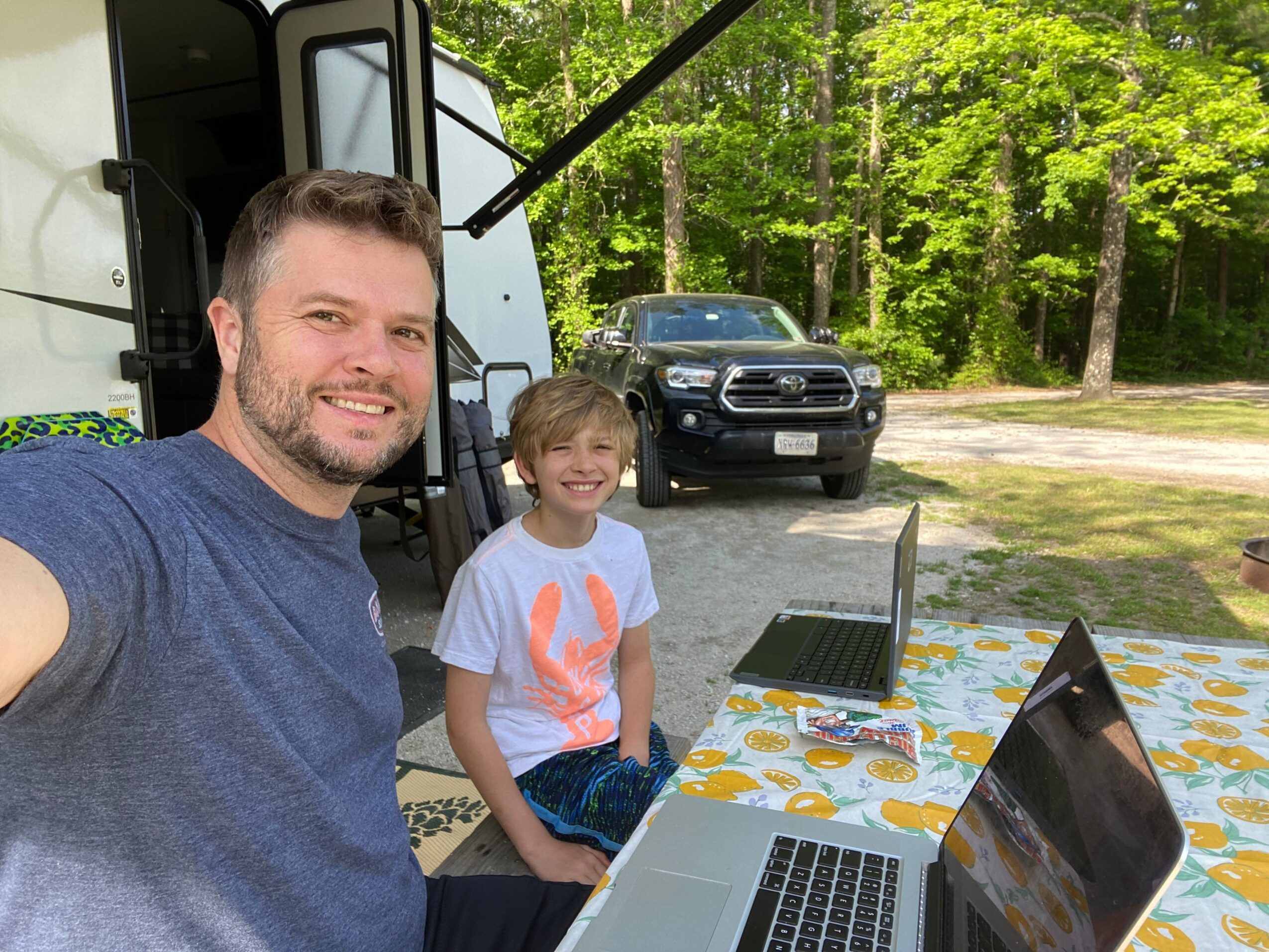 Two children and an adult outdoors near a white vehicle, with trees and a table with a laptop in the background.