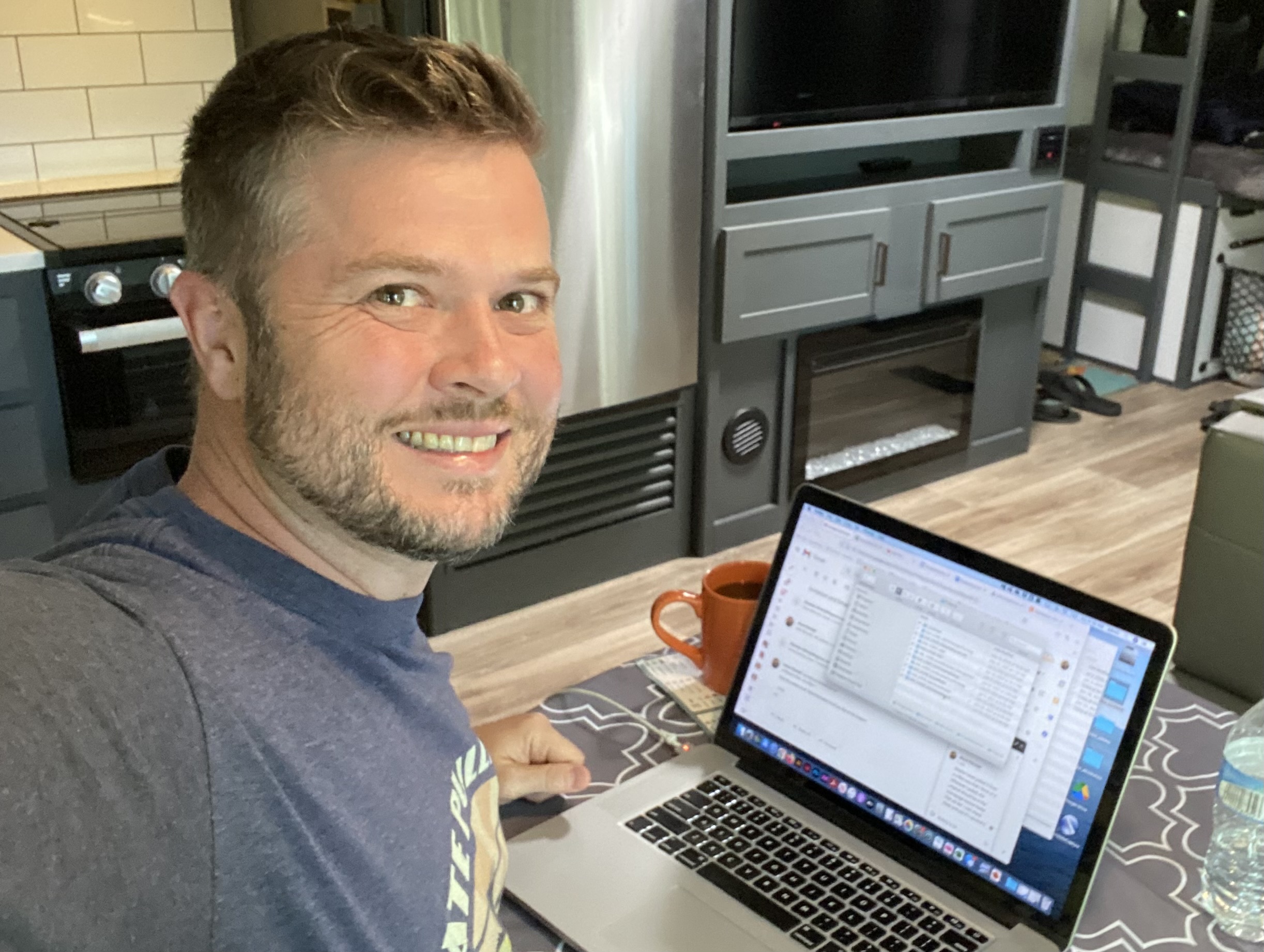 Man with short hair and beard smiling at camera, working on laptop at a table in a kitchen or living room.