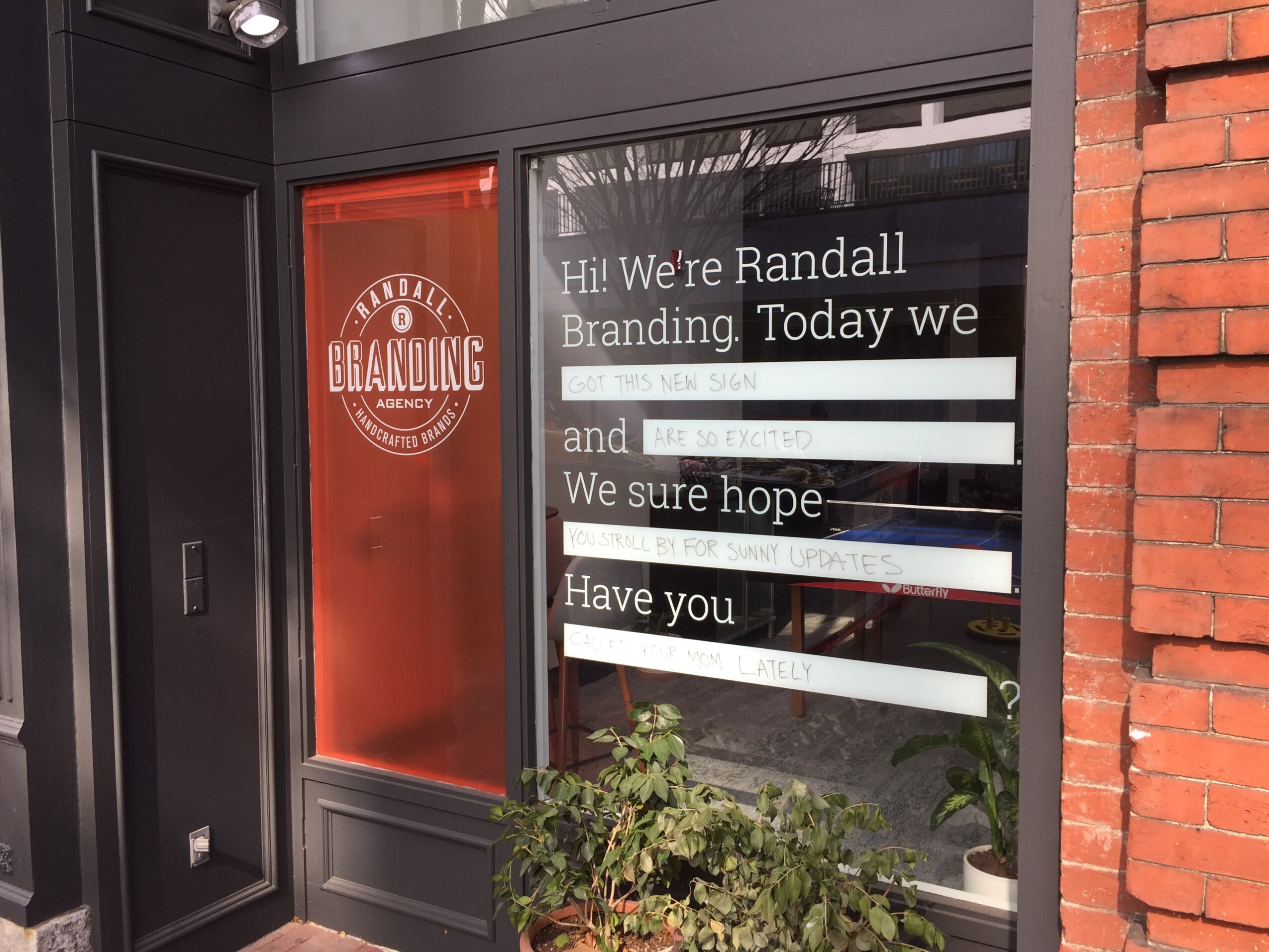Window with text and a plant outside, brick wall on the right, black frame, and a red door with a logo on the left.
