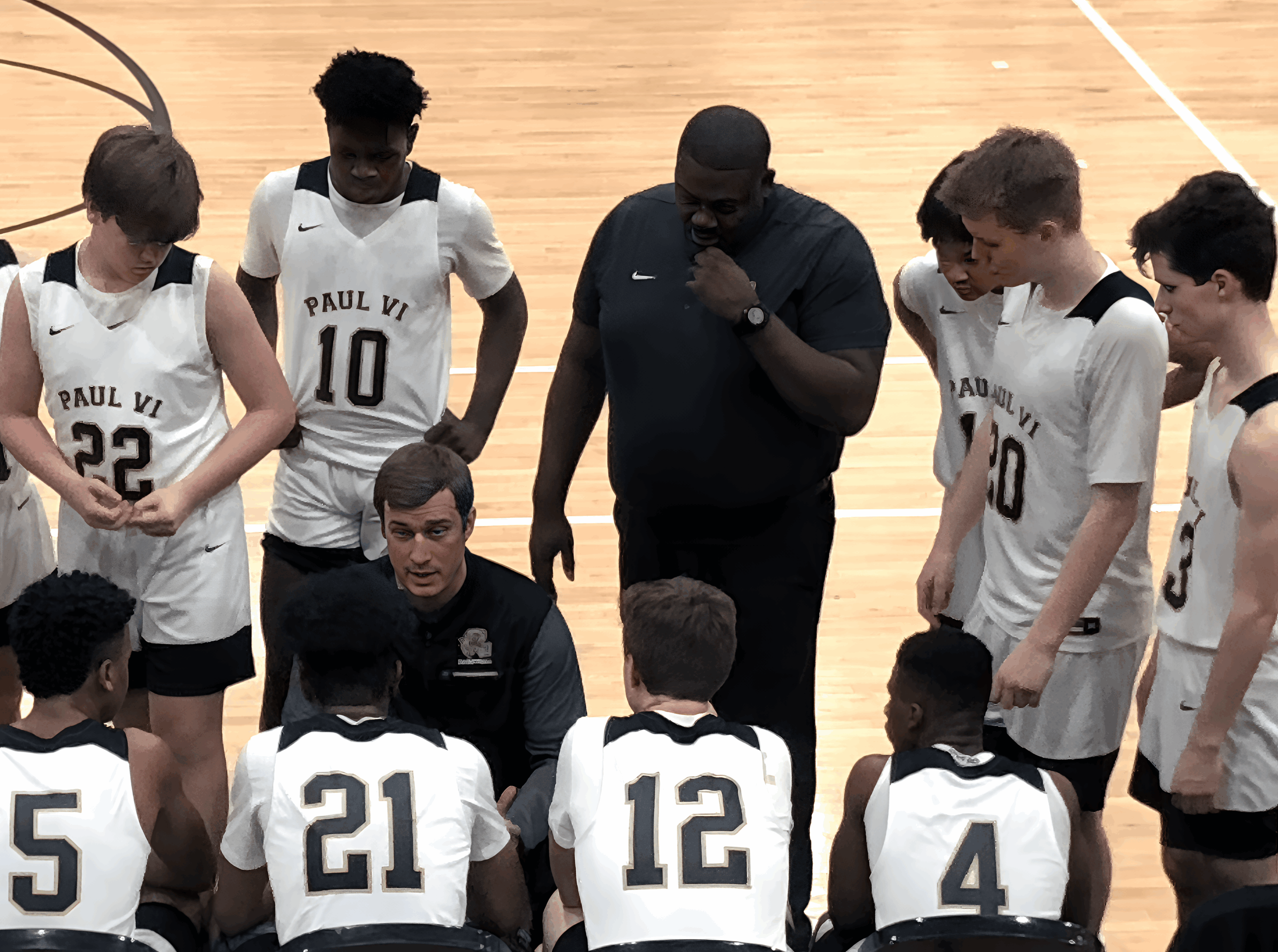 Basketball team huddles around coach on court, players wearing jerseys with numbers, coach giving instructions, hardwood floor visible.
