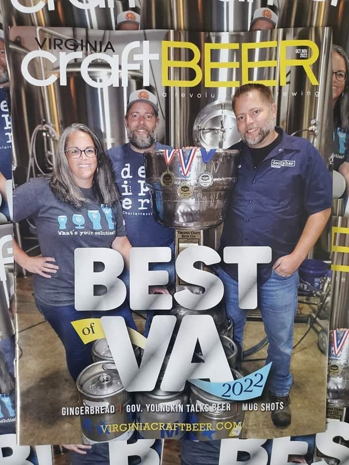 Three people standing in front of beer brewing equipment, holding a large trophy, smiling, with text overlay about Virginia craft beer awards.