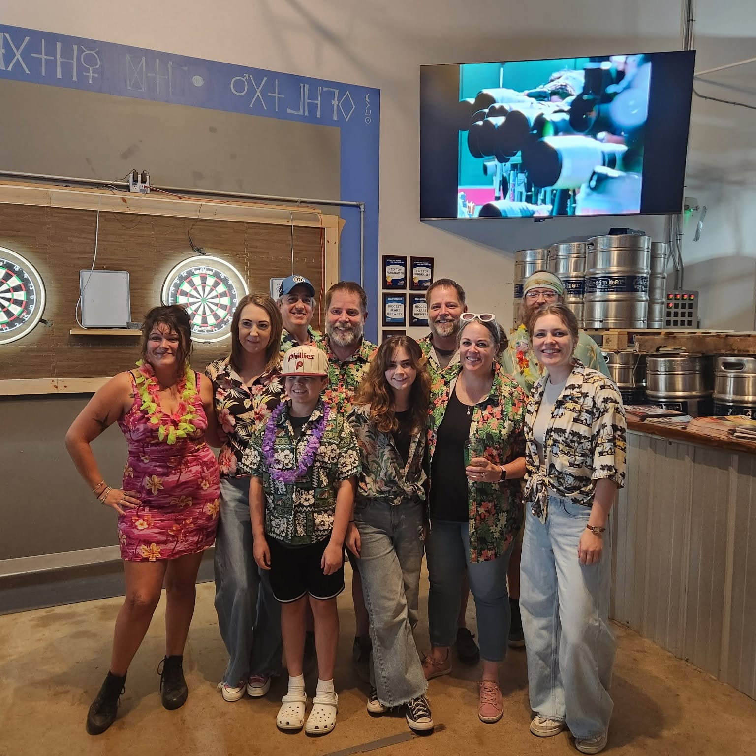 Group of nine people smiling in a bar with dartboards, a TV screen, and bar equipment behind them.