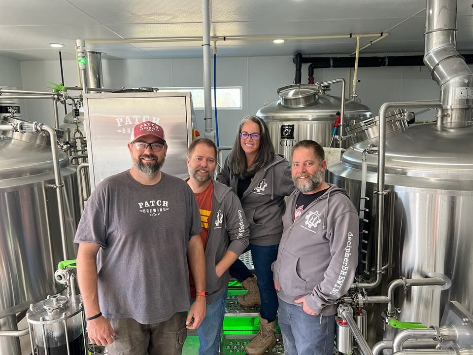 Four people standing in a brewery with large stainless steel tanks and pipes behind them.