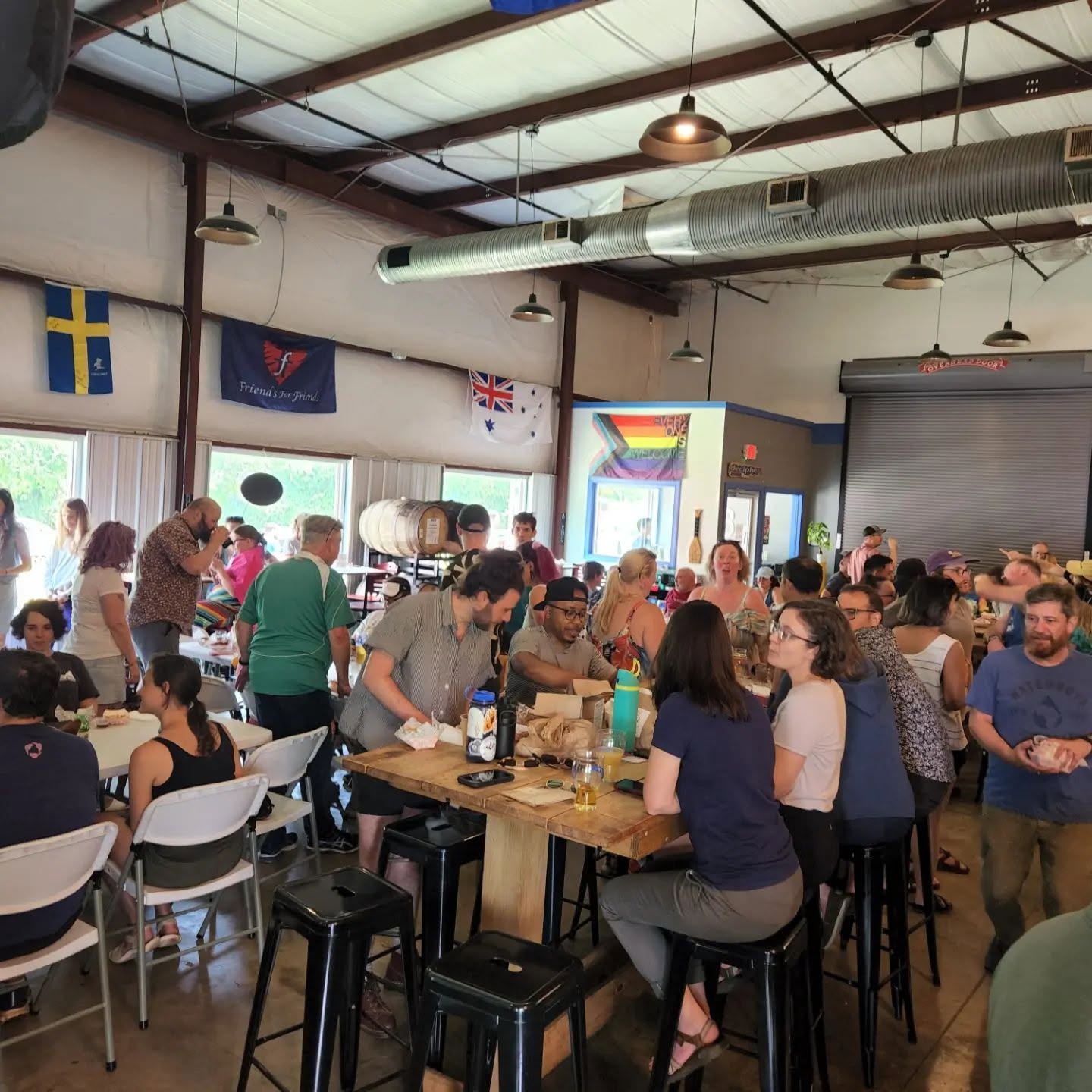 People gather around tables in a large indoor space with flags hanging from the ceiling, some standing and some seated.