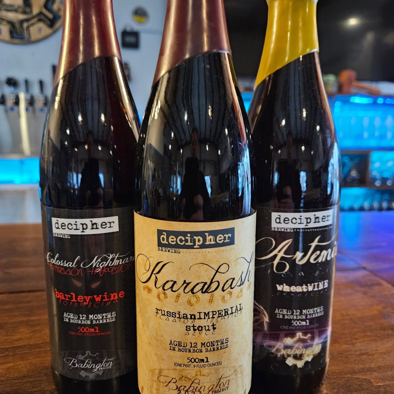 Three bottles of wine on a bar counter, with labels reading 'decipher' and different wine names, in a dimly lit setting.