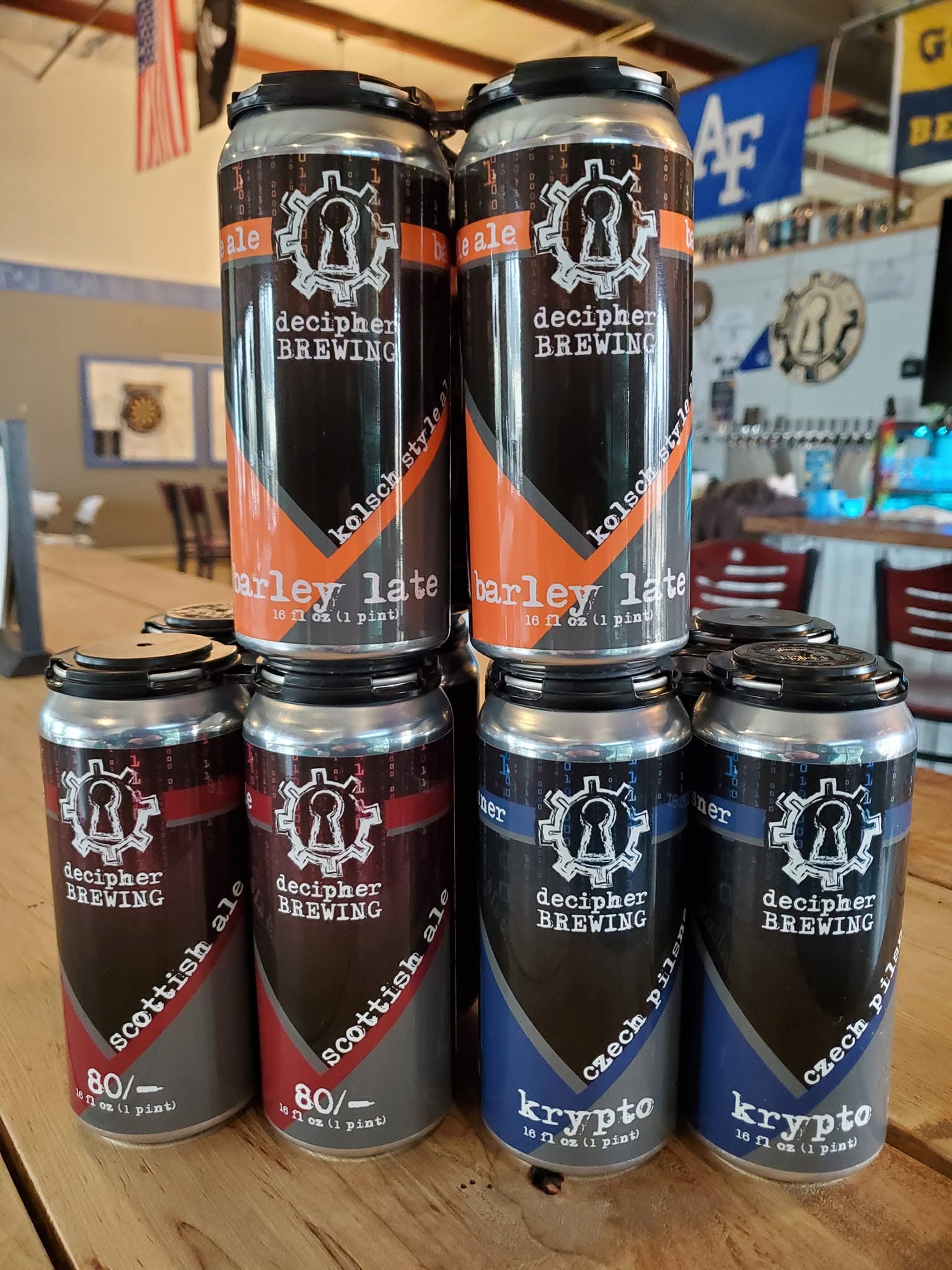 Six cans of Decapitated Brewing beer on a table, with a bar or store background, some cans stacked, colorful labels.